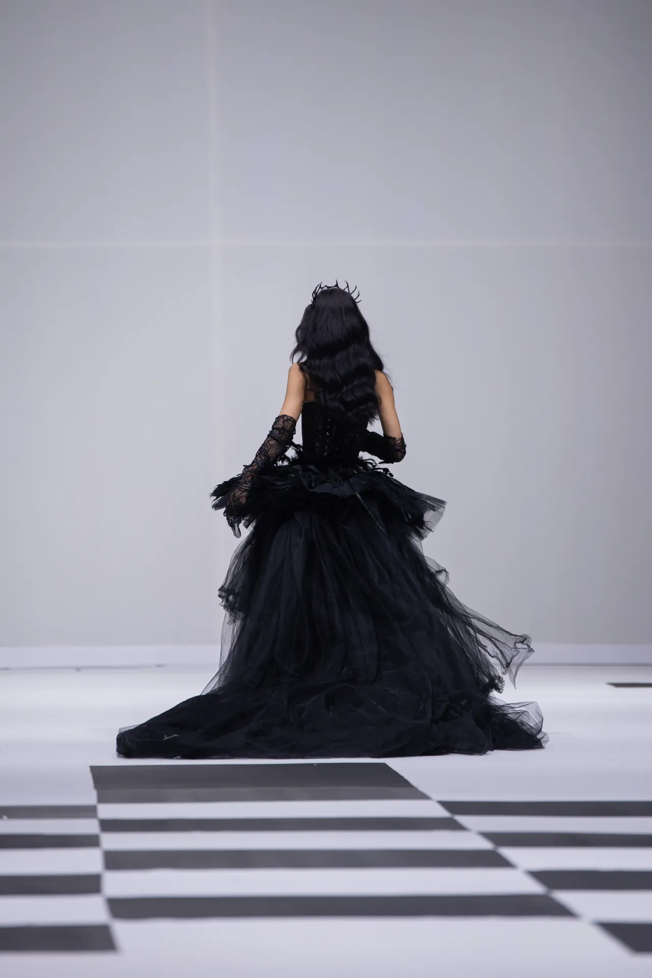 Back view of a model in a dramatic black ball gown with lace gloves, standing on a white runway with a black-and-white checkerboard floor.
