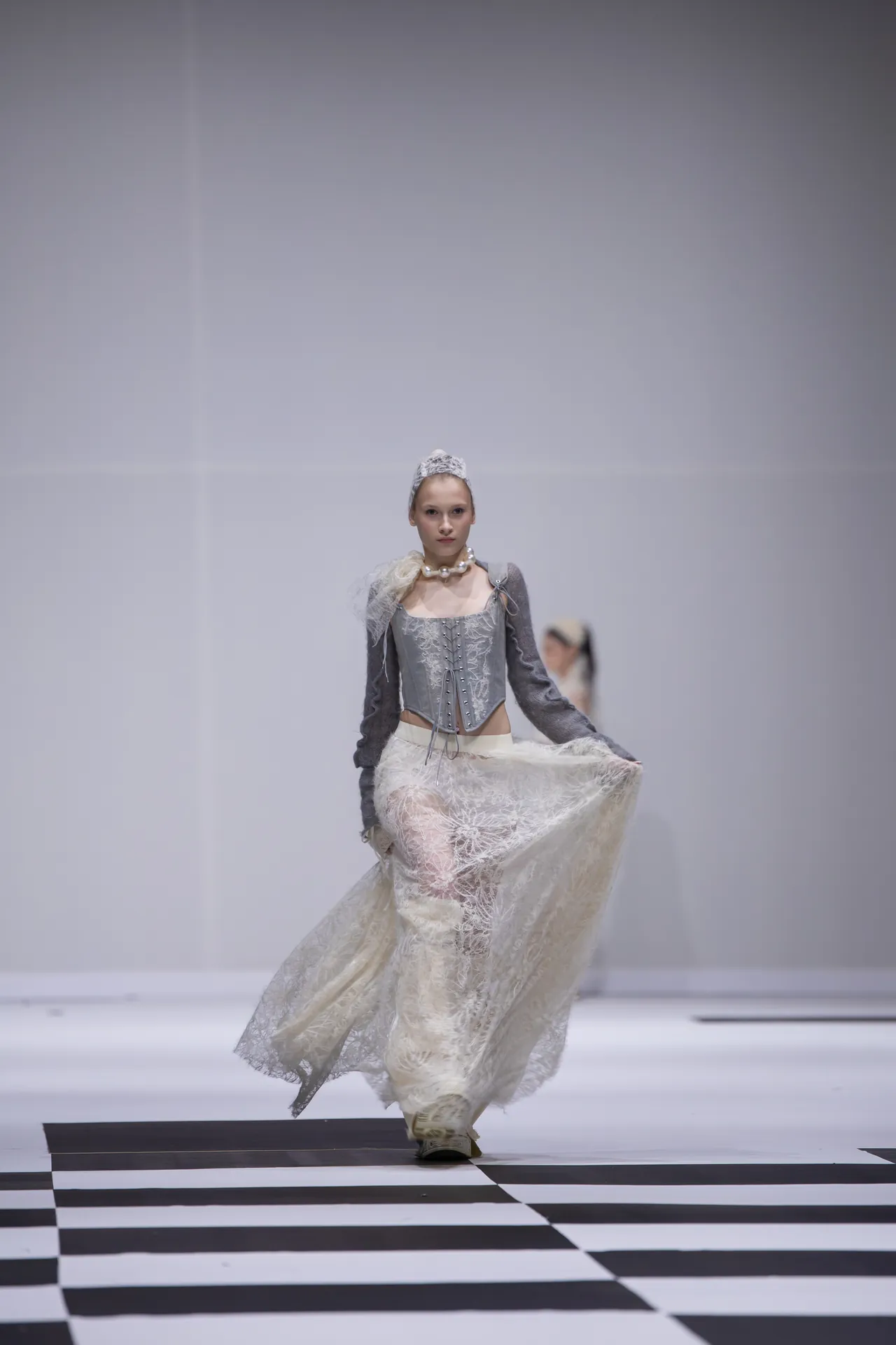 Model walking a runway in a gray corset top and lace skirt, with a pearl necklace and headpiece.