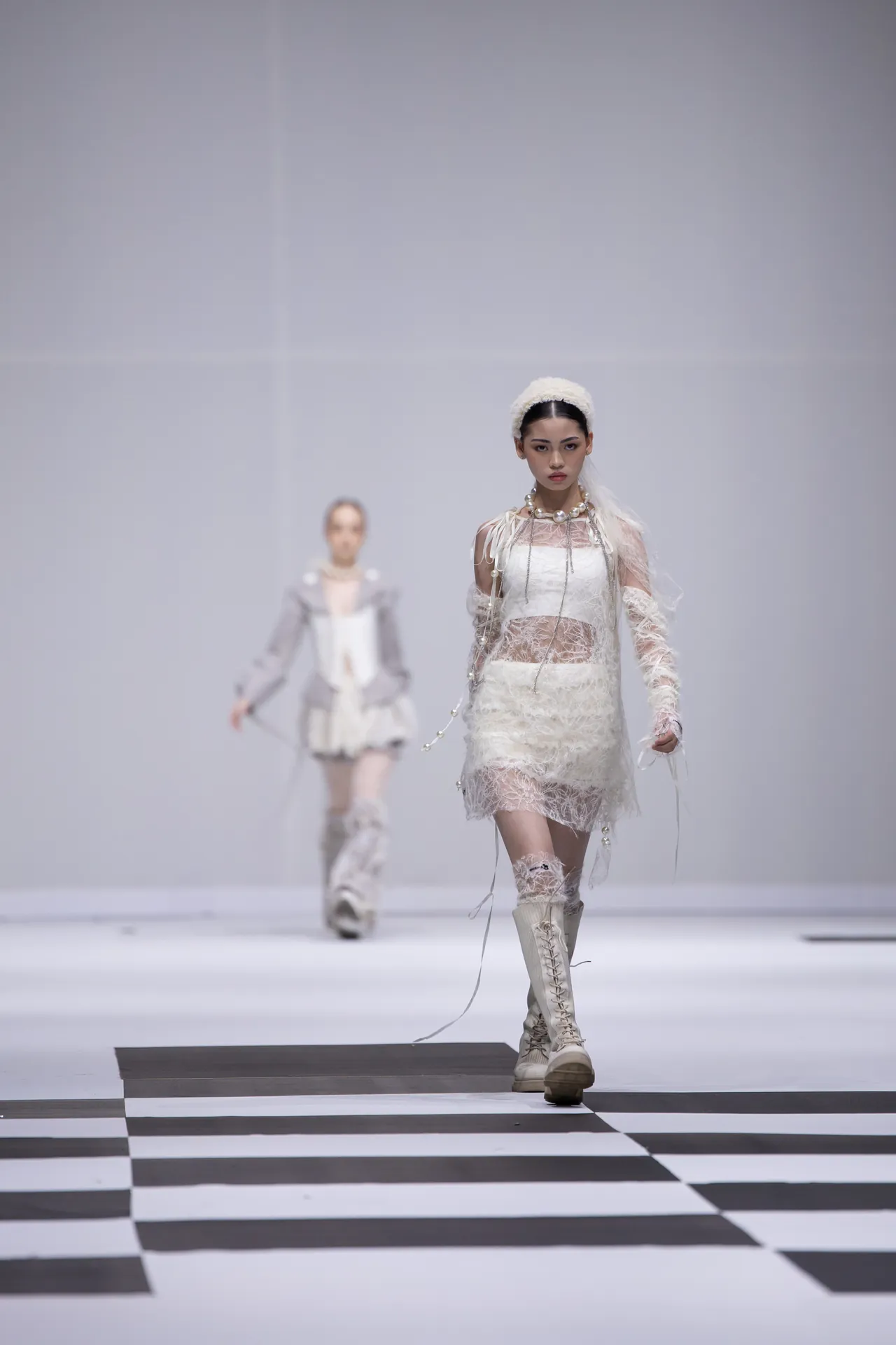 Model walks a white lace dress and knee-high lace-up boots on a bold black-and-white checkered runway.