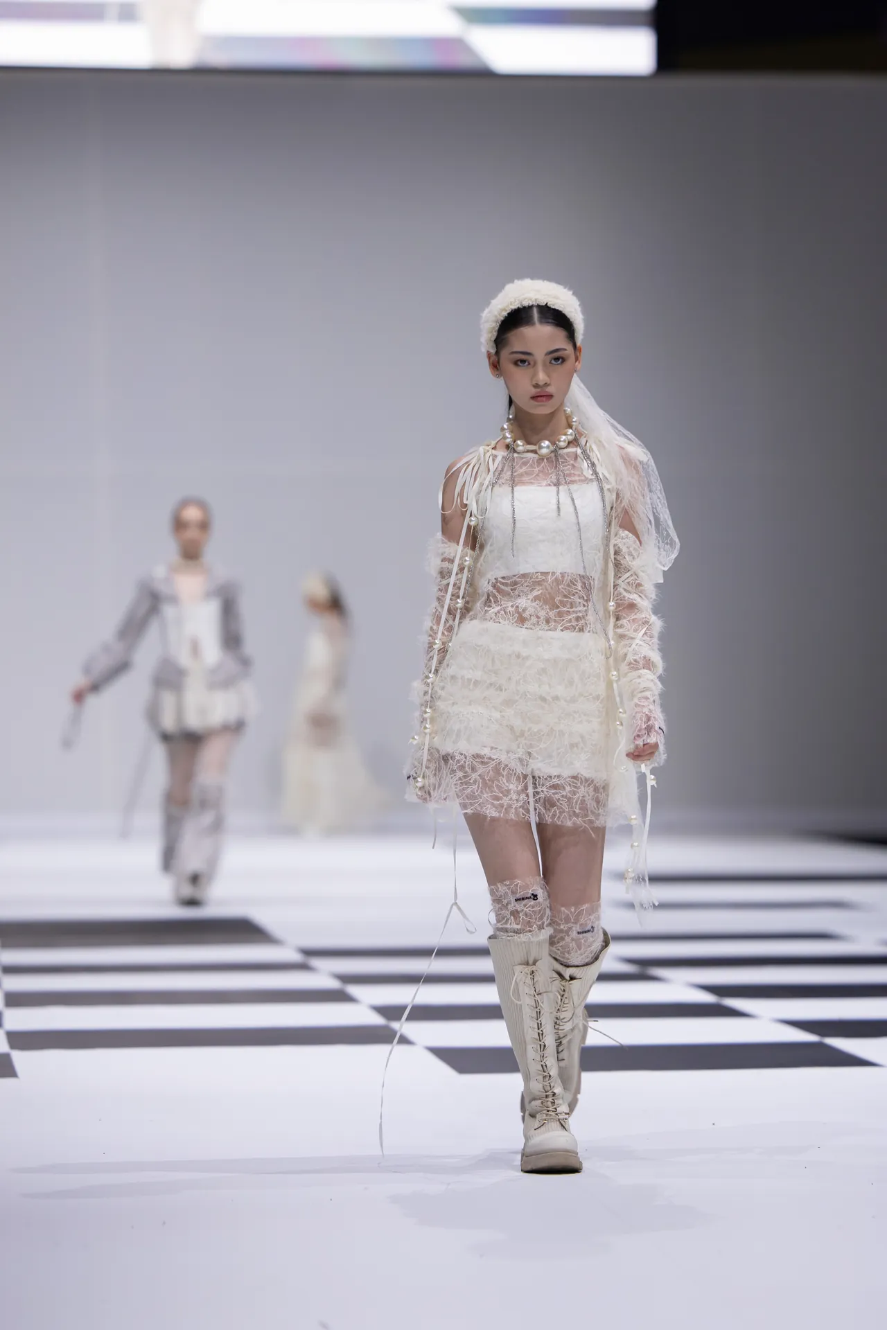 Model walks the runway in a white lace dress with a chunky pearl necklace and knee-high lace-up boots on a striped catwalk.