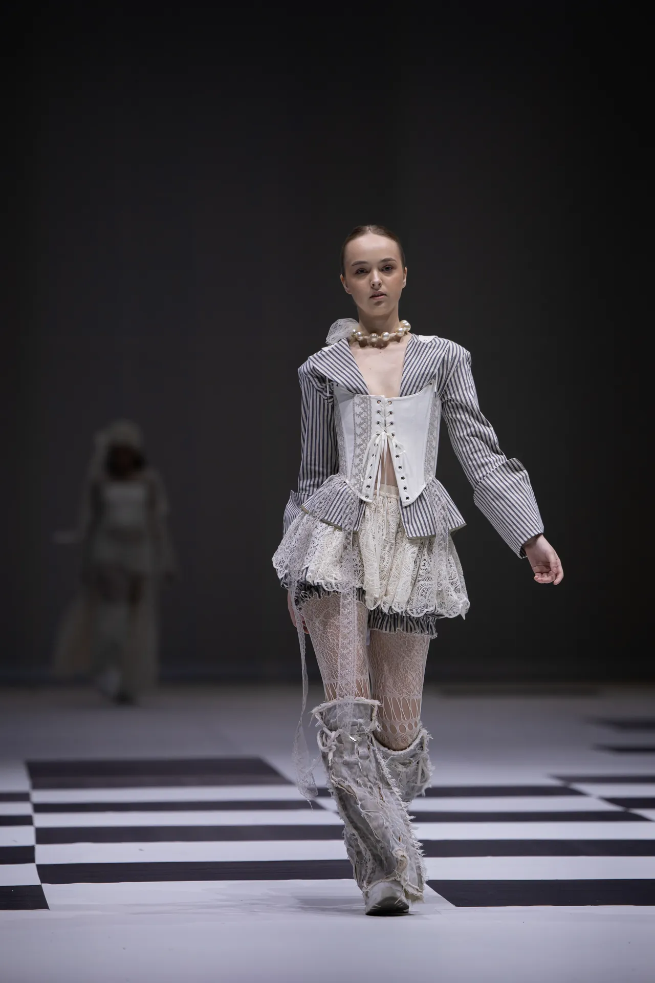 Model walking a fashion runway in a Victorian-inspired striped jacket, lace skirt, corset, and pearl necklace on a black-and-white checkerboard stage.