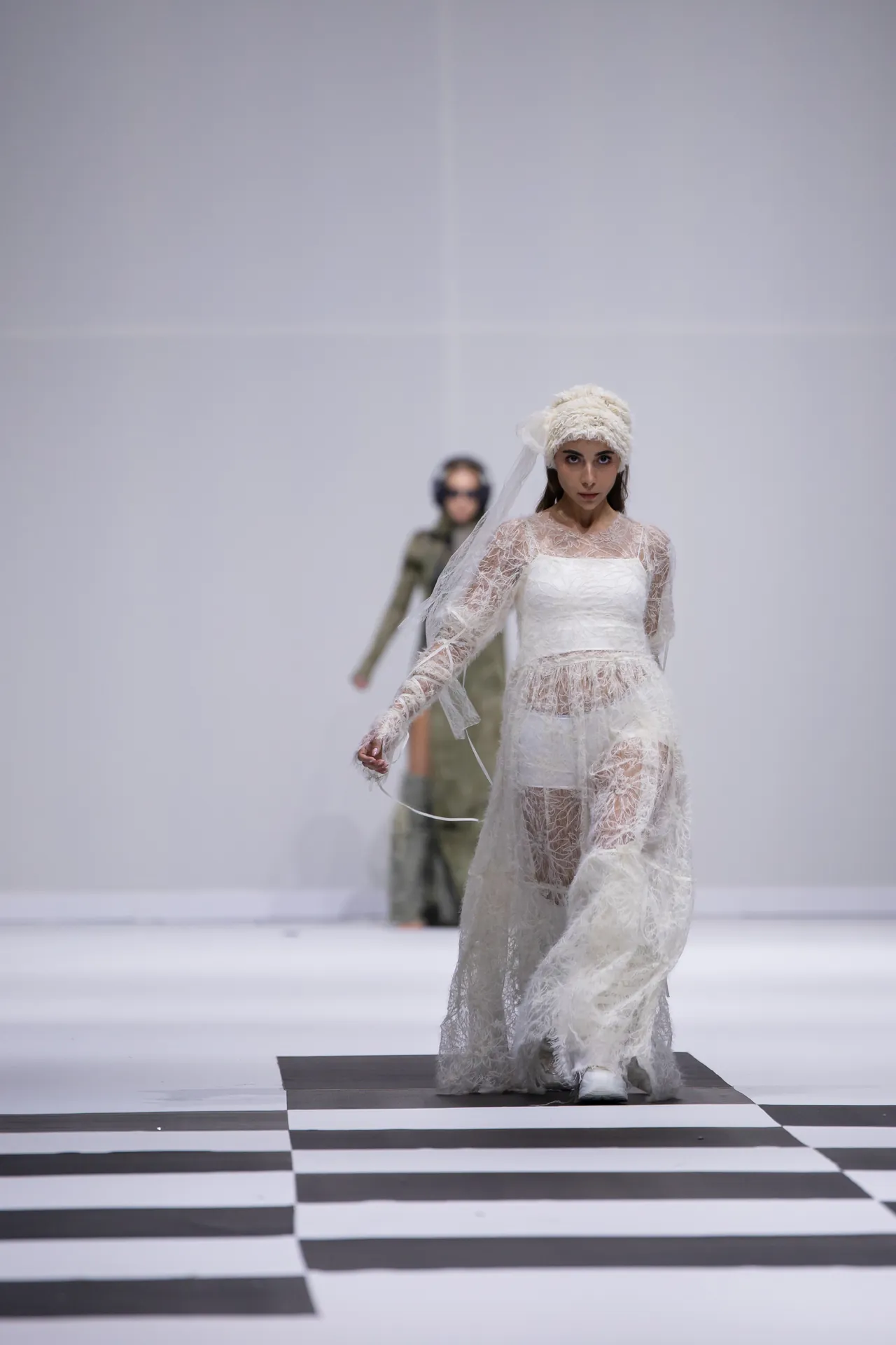 Female model walking a fashion runway in a white lace wedding dress with a veil and headpiece; black-and-white checkered floor and a minimal backdrop.