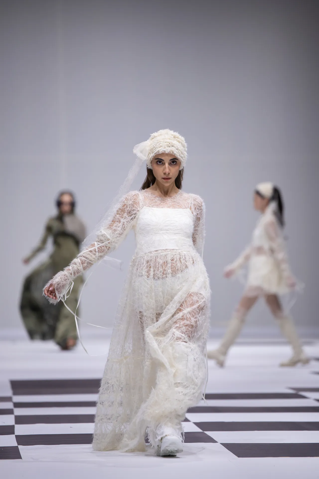 Model walks a white lace wedding-inspired gown with long sleeves and a sheer veil on a checkered runway.