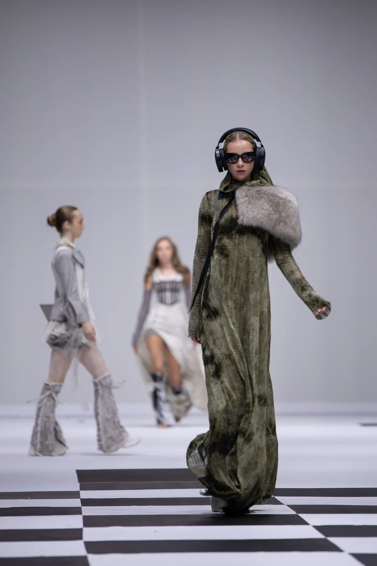 Model walks the runway in a long green gown with a fur stole, wearing large black headphones and sunglasses.