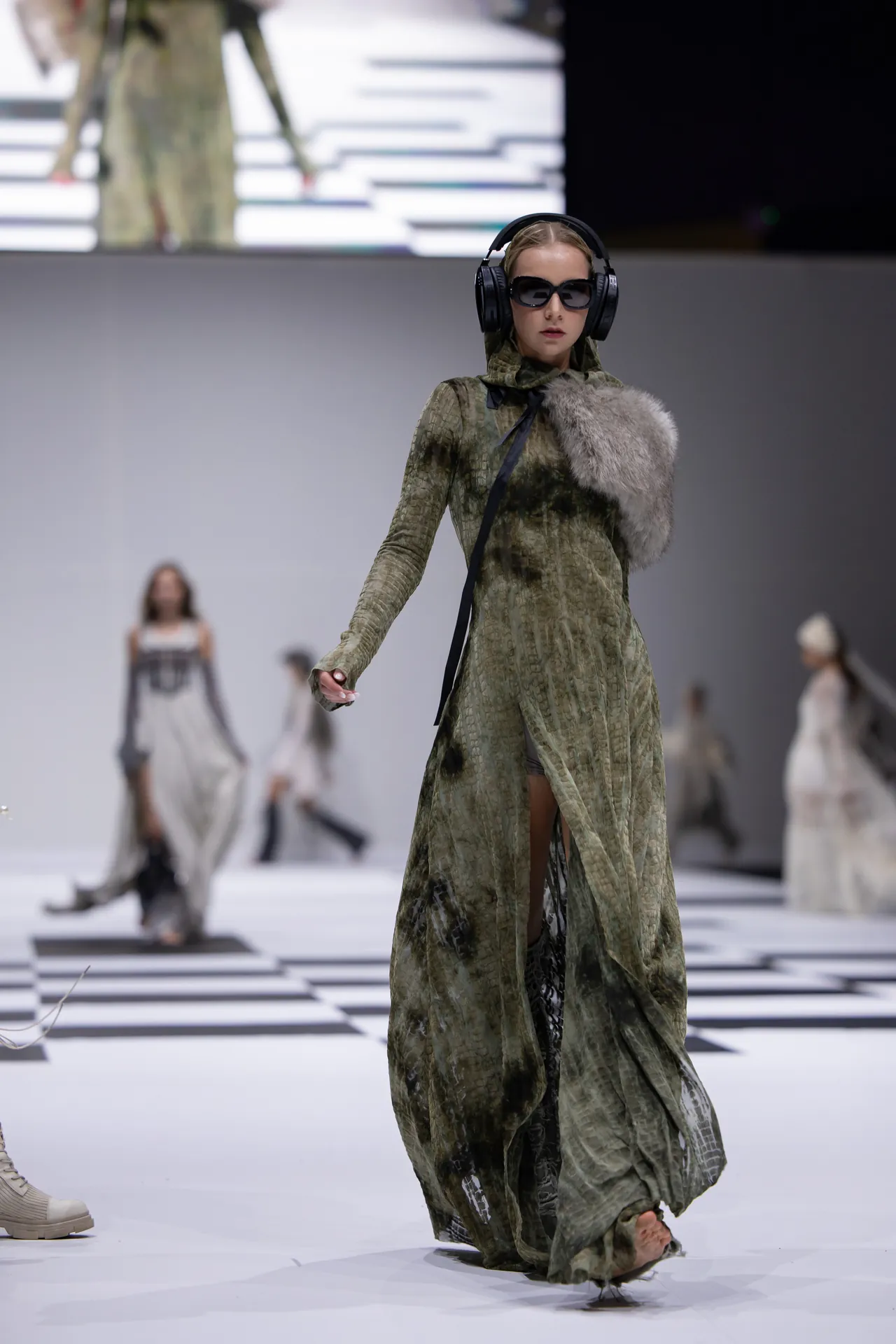 Runway model in a green camouflage gown with a gray fur stole, wearing large black headphones and sunglasses as she walks the catwalk.