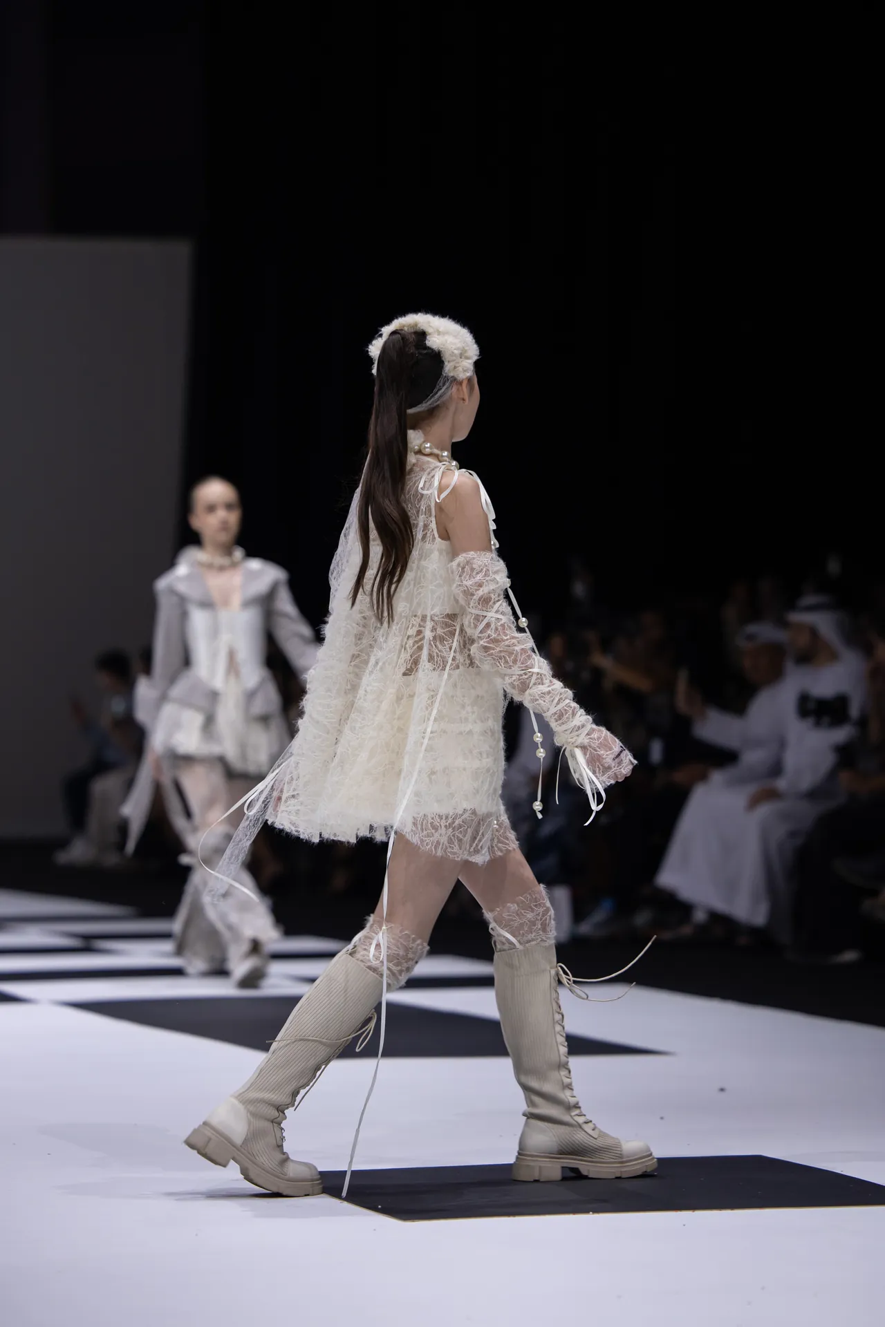 Model walking the runway in a sheer ivory lace outfit with thigh-high boots and a fluffy headpiece.