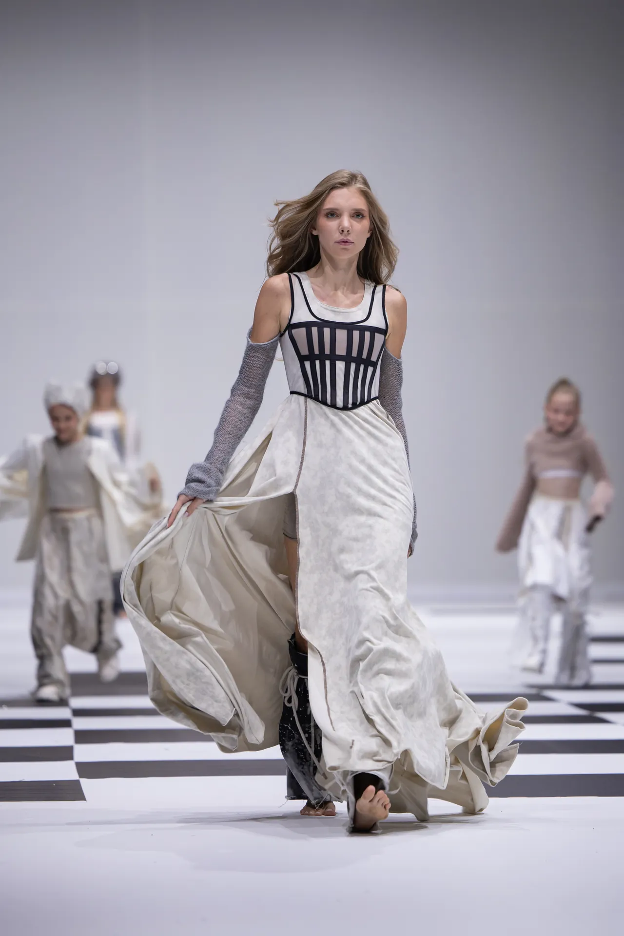 Model walking a fashion runway in a white gown with a black corset bodice and flowing sleeves on a black-and-white checkered floor, mid-stride.