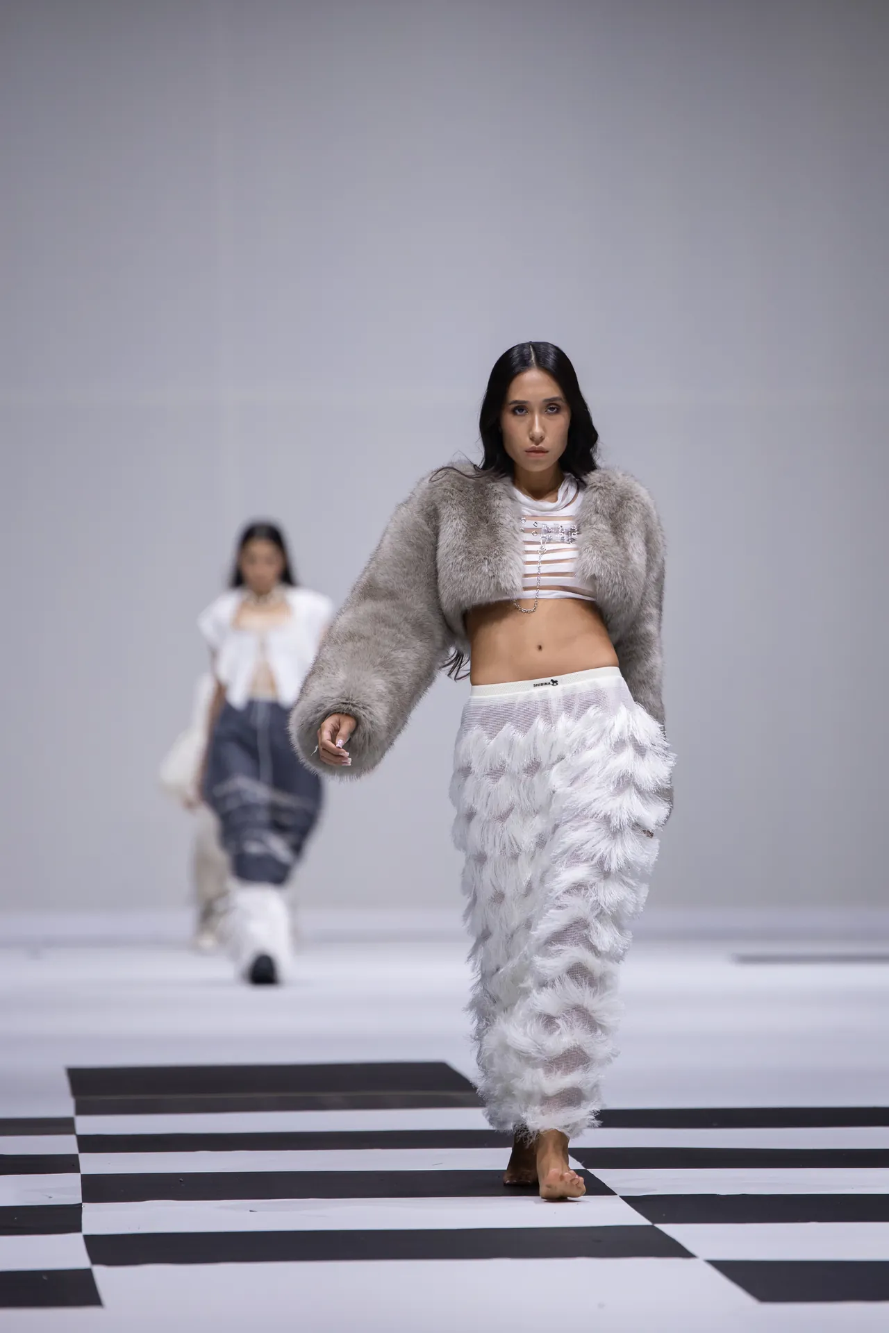Model walks a fashion runway in a light gray fur coat and white feathered pants on a black-and-white checkerboard catwalk.]","Runway scene with a female model wearing a gray fur coat and feathered white pants, approaching the camera."","Fashion show moment: model struts down a checkerboard catwalk in a fur coat and feathered pants."","Minimalist runway shot of a model in a gray fur coat and white feathered pants, on a high-contrast catwalk."","A model models a bold winter ensemble on a black-and-white checkerboard runway.