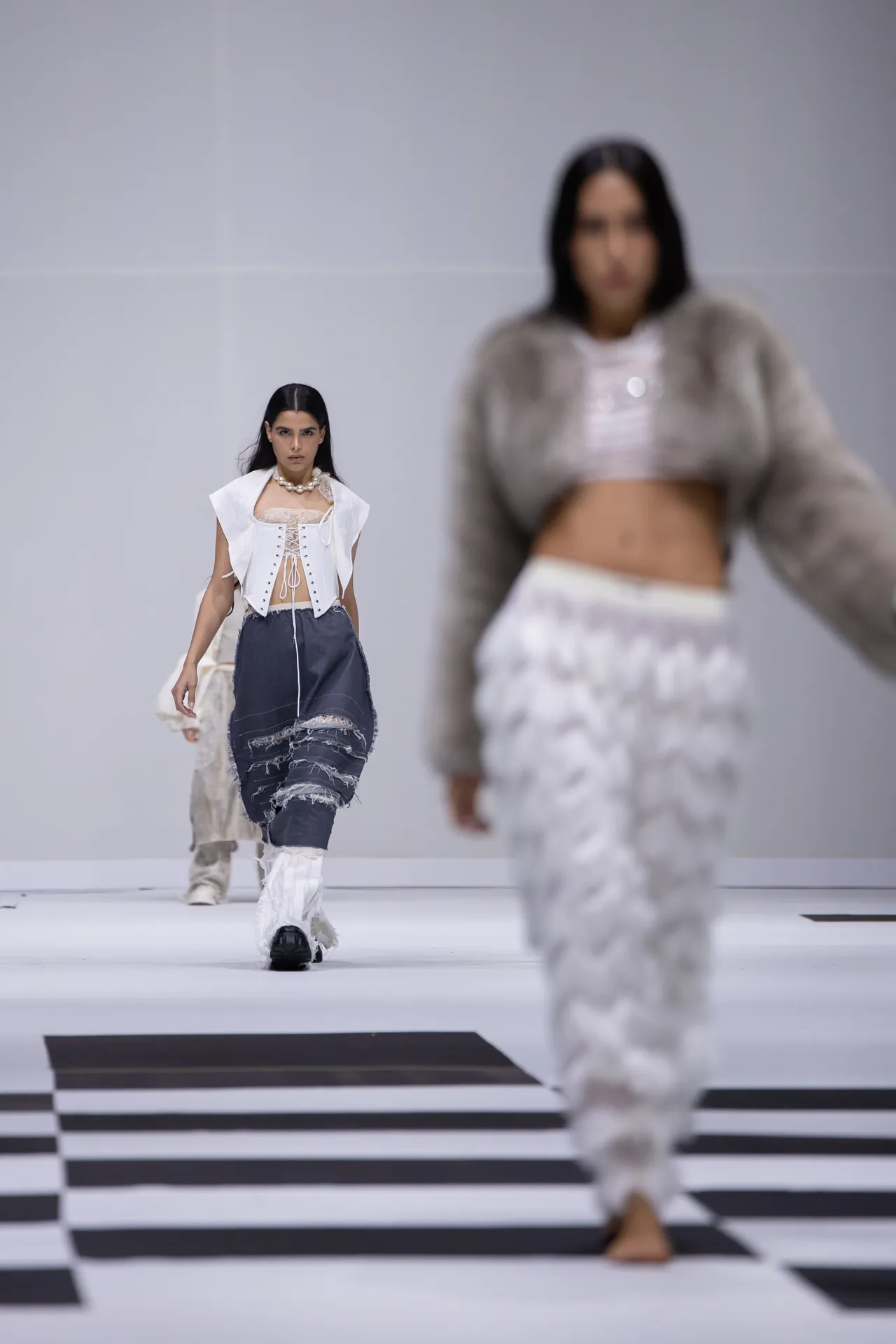 Model on a fashion runway wearing a white corset-style top and distressed navy skirt; black-and-white striped floor, foreground model blurred.