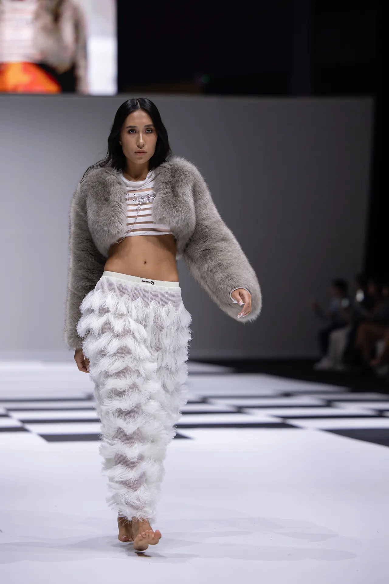 Female model walks the runway in a light gray faux fur coat over a white cropped top and a fluffy white feathered skirt/pants ensemble, on a black-and-white checkered runway.