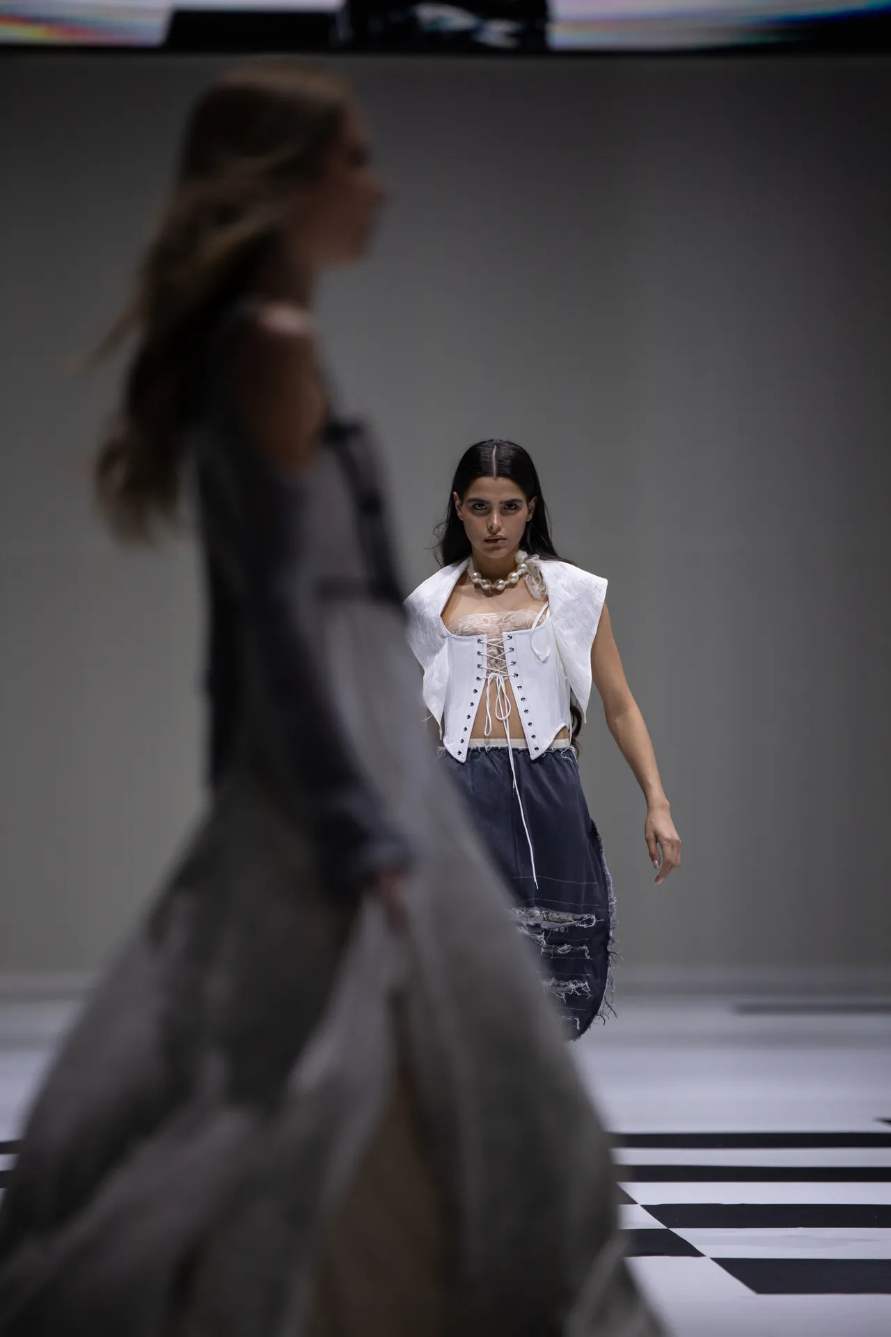 Model walking a fashion runway in a white corseted top with lace-up front, layered over a sheer blouse, paired with a ripped denim skirt and a pearl necklace.