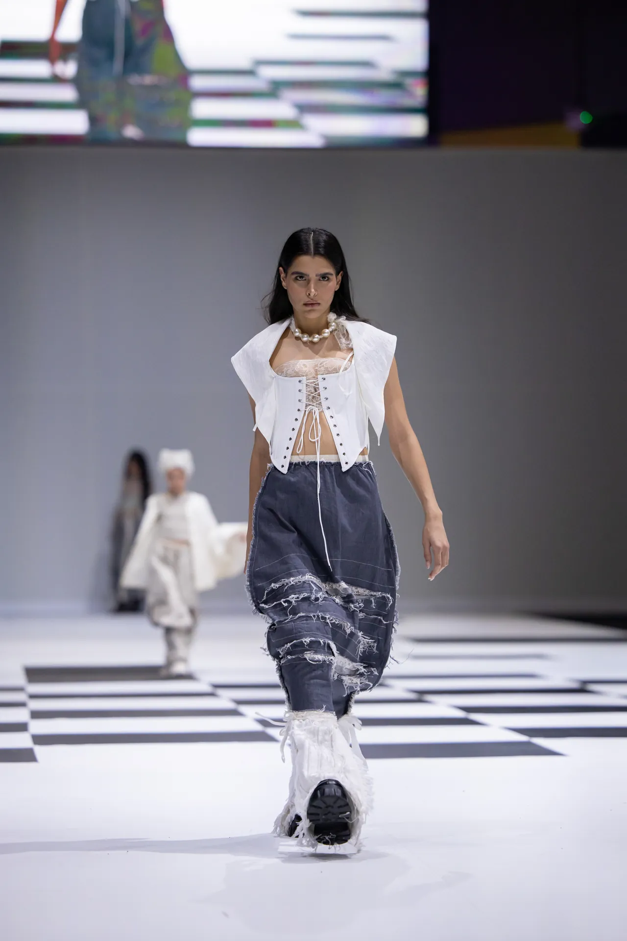 Model walks a fashion runway in a white corset top with lace, a white vest, ripped denim pants, chunky white fur boots, and a pearl necklace on a black-and-white checkered floor.