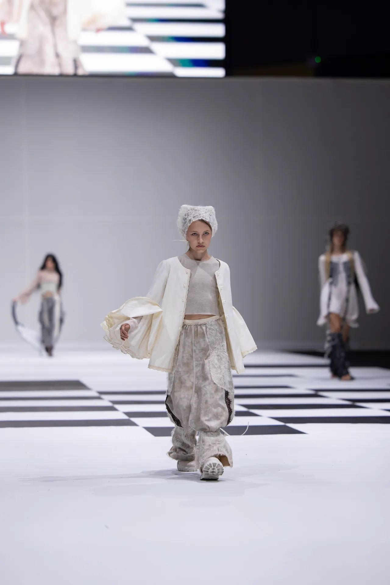Young child model walks down a white runway in a cream, textured outfit with a fur hat, against a black-and-white checkered floor. Other models appear blurred in the background.