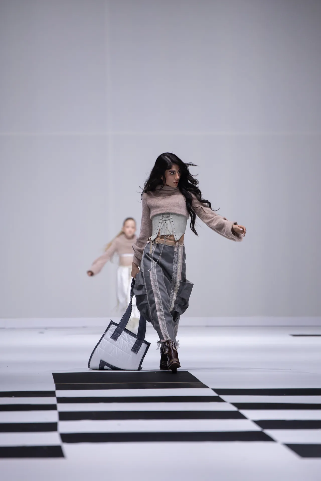 Model walking down a monochrome striped runway in a beige sweater and corset, carrying a large white tote bag.