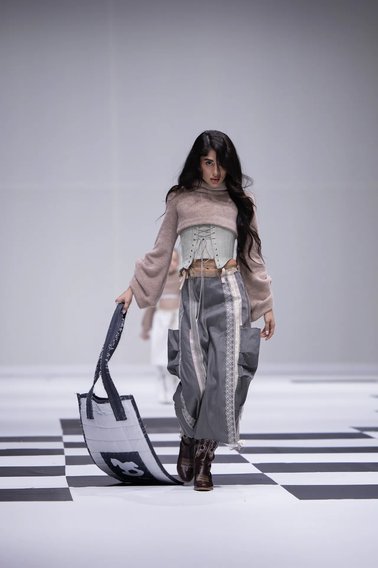 Model walks a fashion runway on a black-and-white checkered floor, wearing a beige knit top with a corset belt and gray patterned pants, carrying a white quilted bag with dark straps.