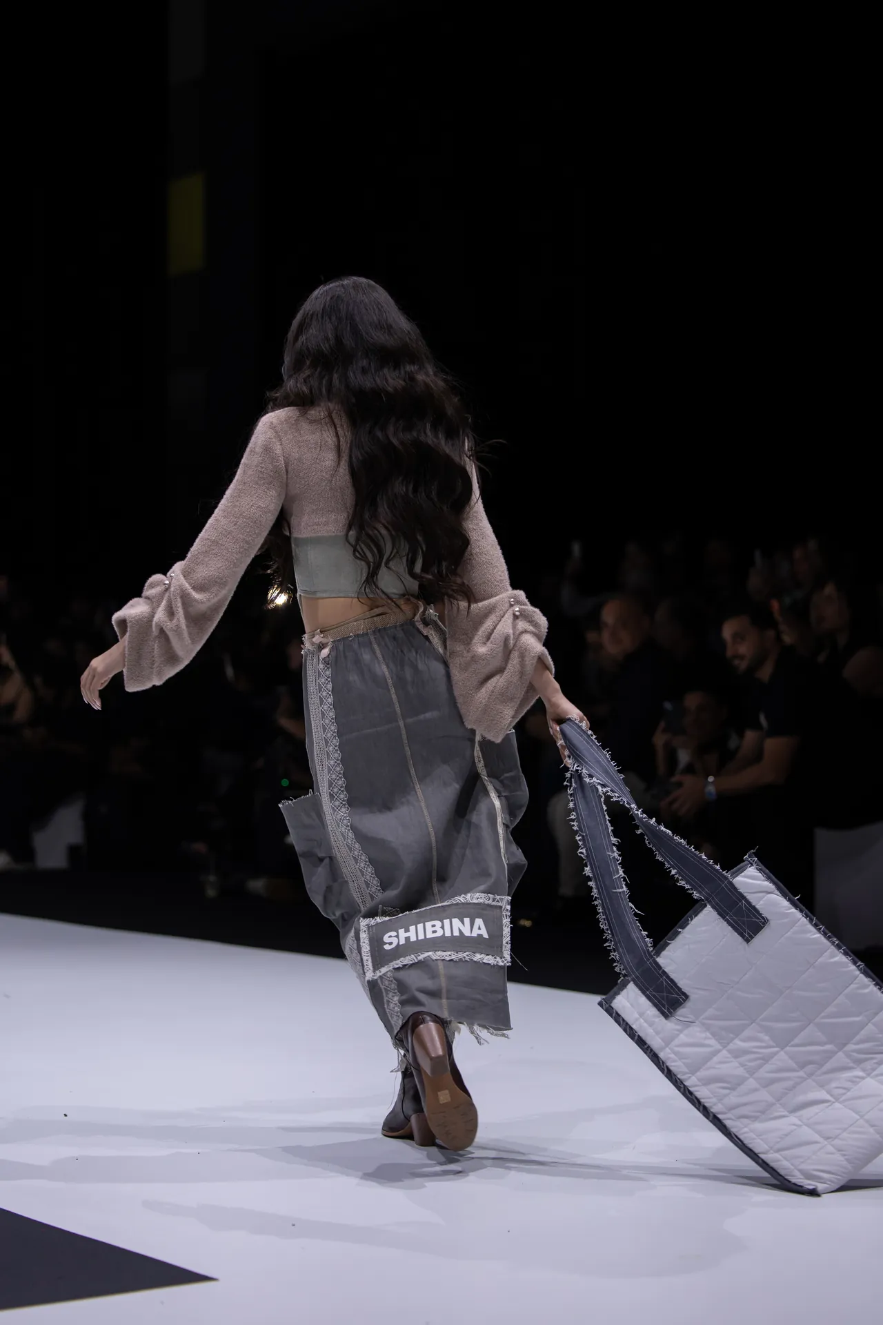 Back view of a fashion model walking the runway, wearing a beige textured top and gray patchwork pants with a SHIBINA label, carrying a large quilted tote bag.