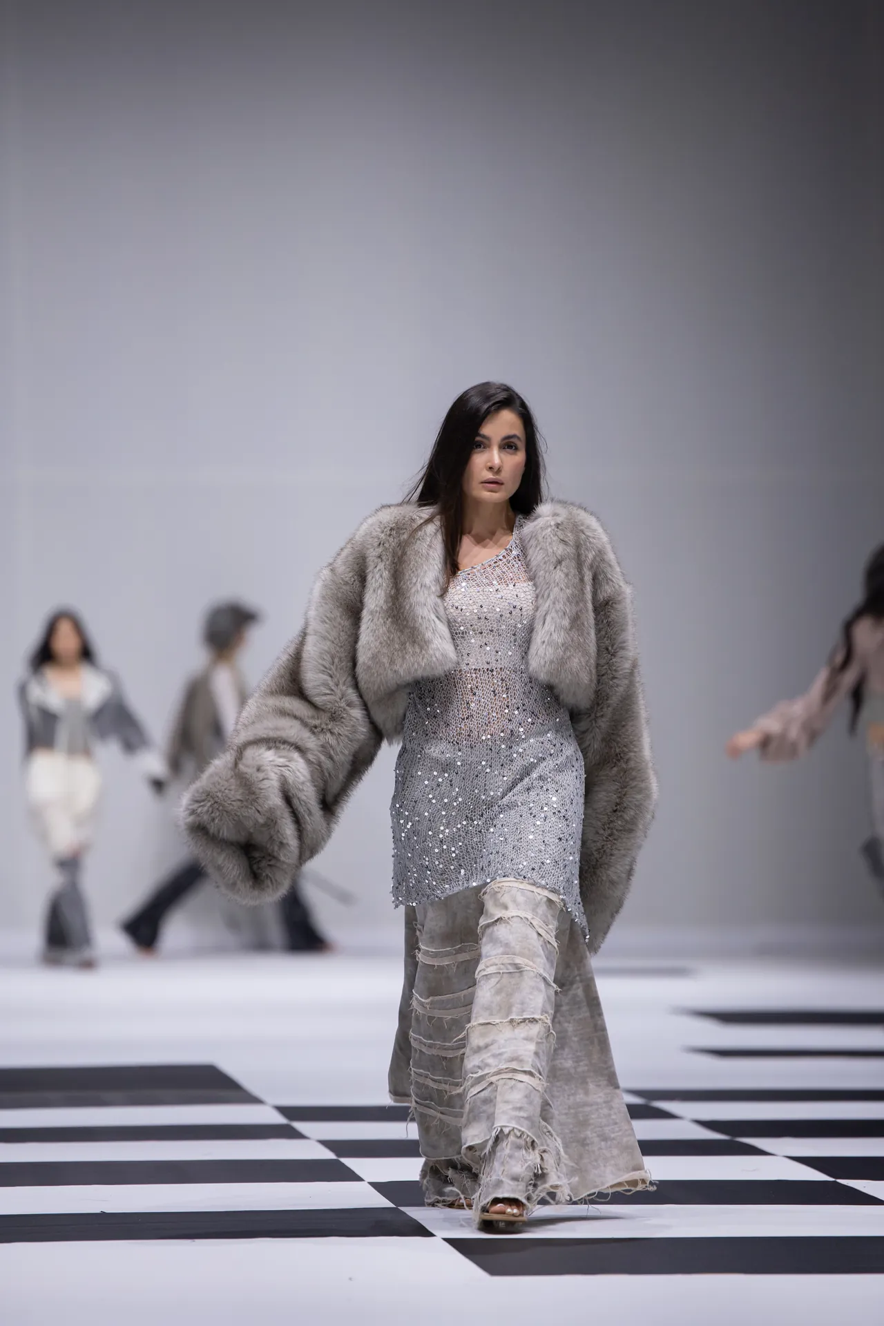 Model walks a fashion runway in a silver sequined dress and gray fur coat against a black-and-white checkered floor.