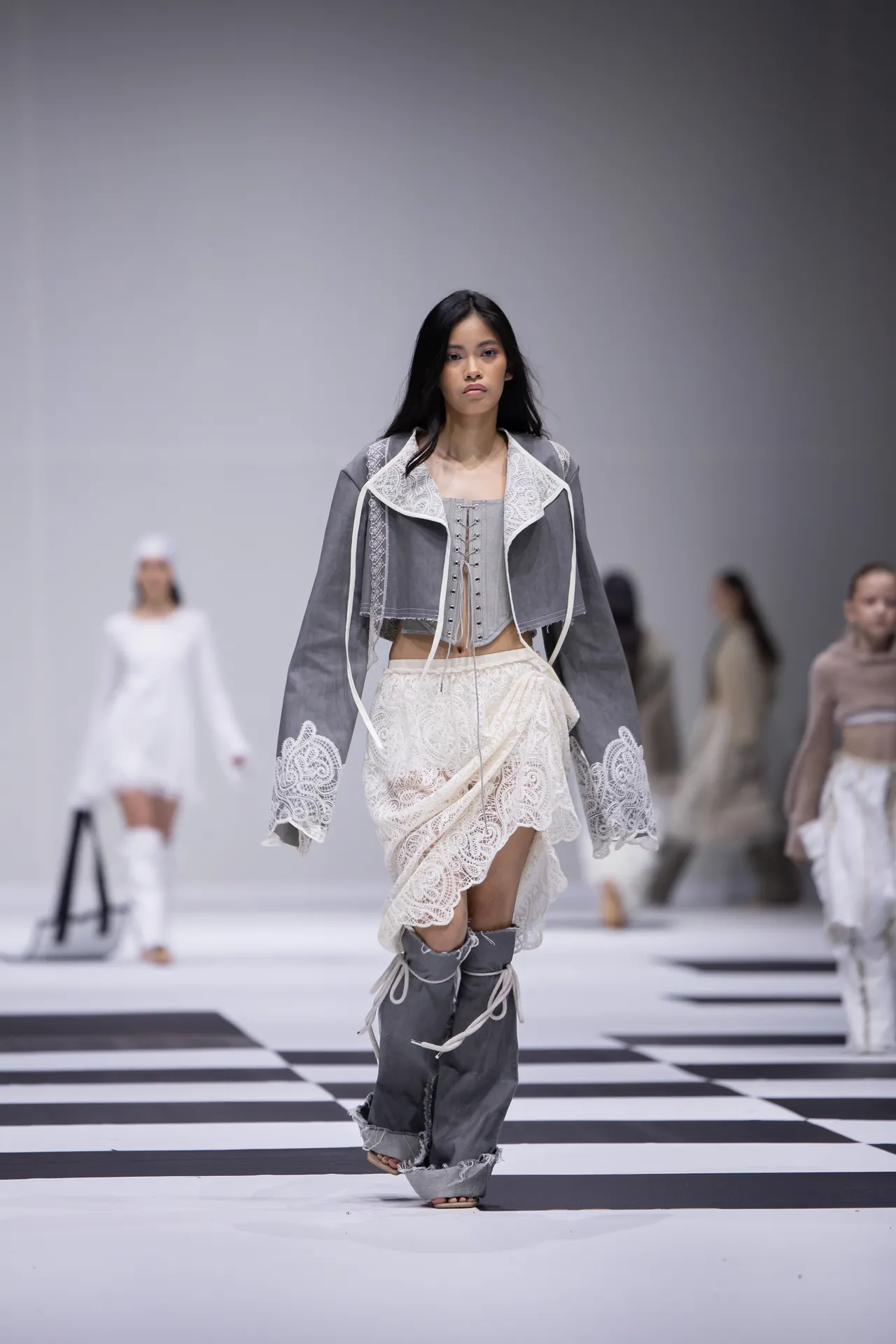 Model walks the runway in a gray lace-trimmed jacket over a corset top and white lace skirt.