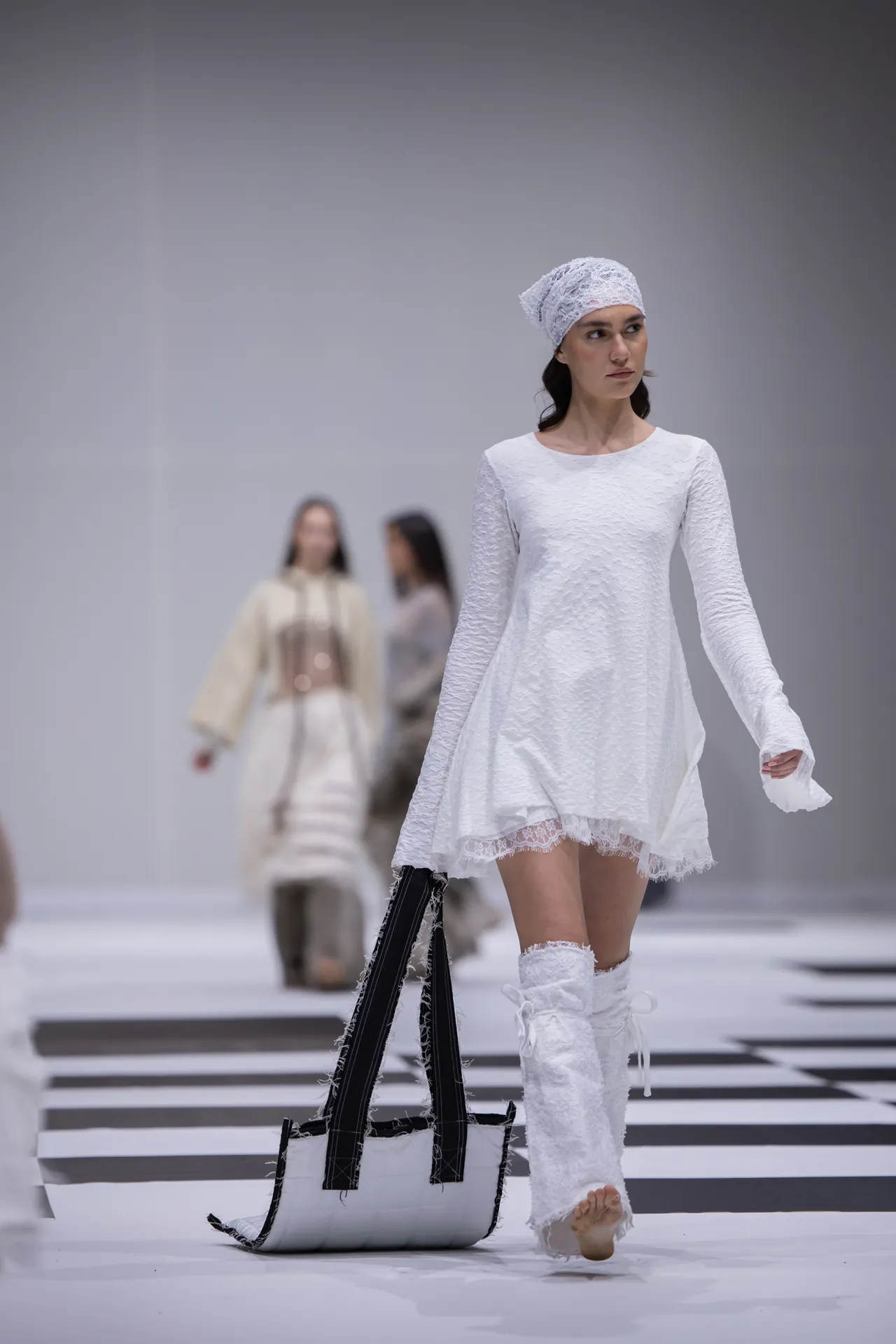 Model walks down a runway in a white lace dress with a matching headscarf, carrying a large black-and-white bag on the floor beside her.