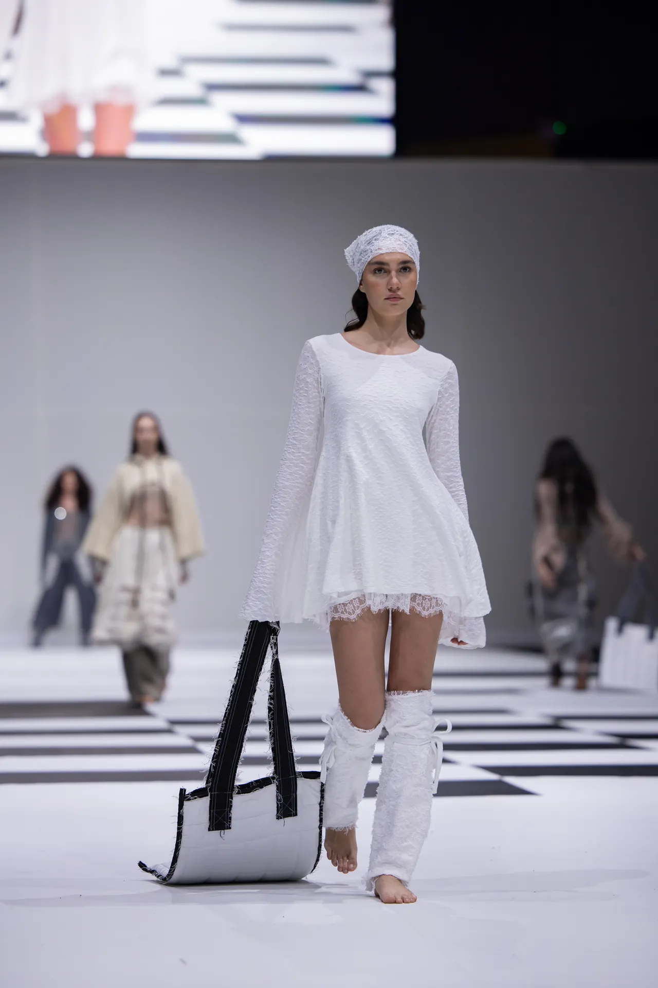 Runway model in a white lace dress and head wrap, walking barefoot with a large black-and-white tote on the floor nearby.