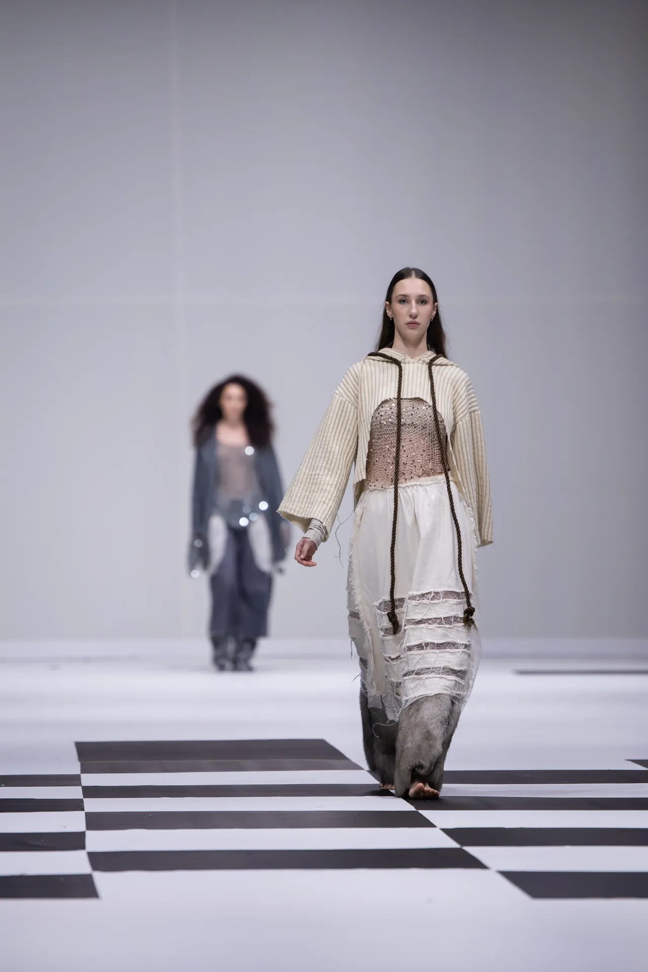 Model walks a black-and-white checkerboard runway in a cream knit top over a lace dress, centered on the catwalk.