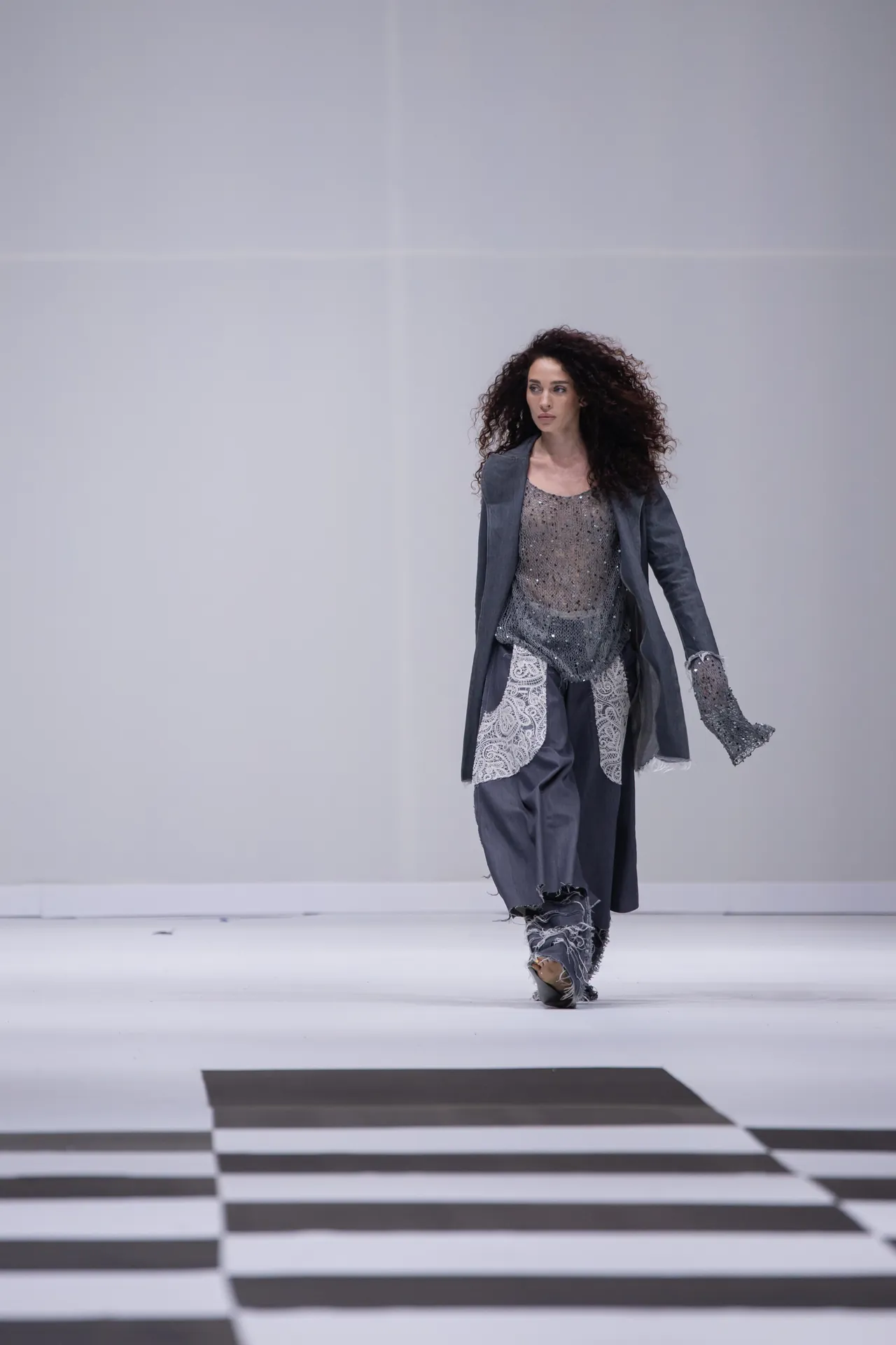 Model walks the runway wearing a metallic gray sequin top, matching draped coat, and lace-trimmed pants on a black-and-white checkered catwalk.