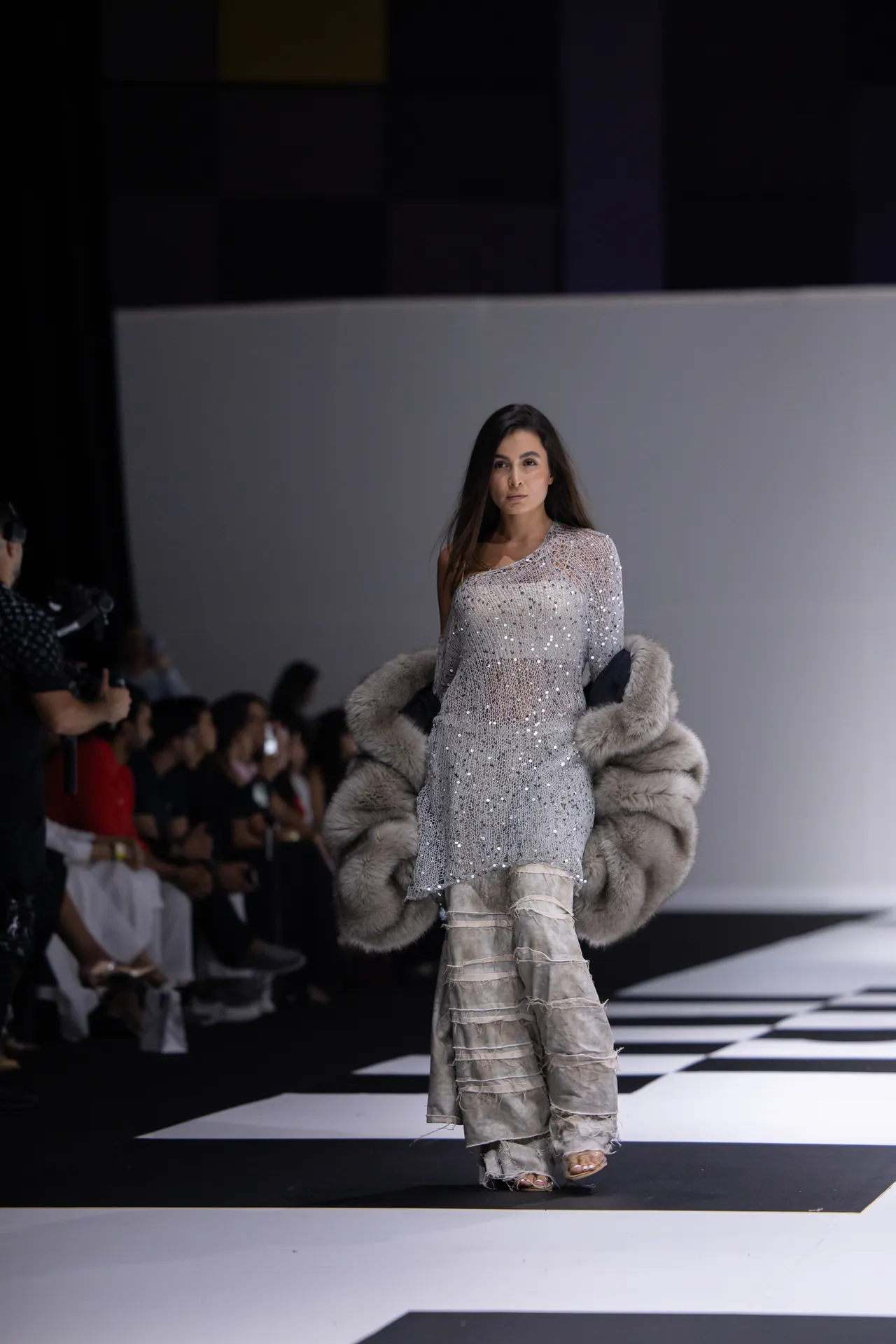 Model walking a fashion runway in a silver sequined dress with a large fur stole, against a black-and-white checkered floor and an audience in the background.