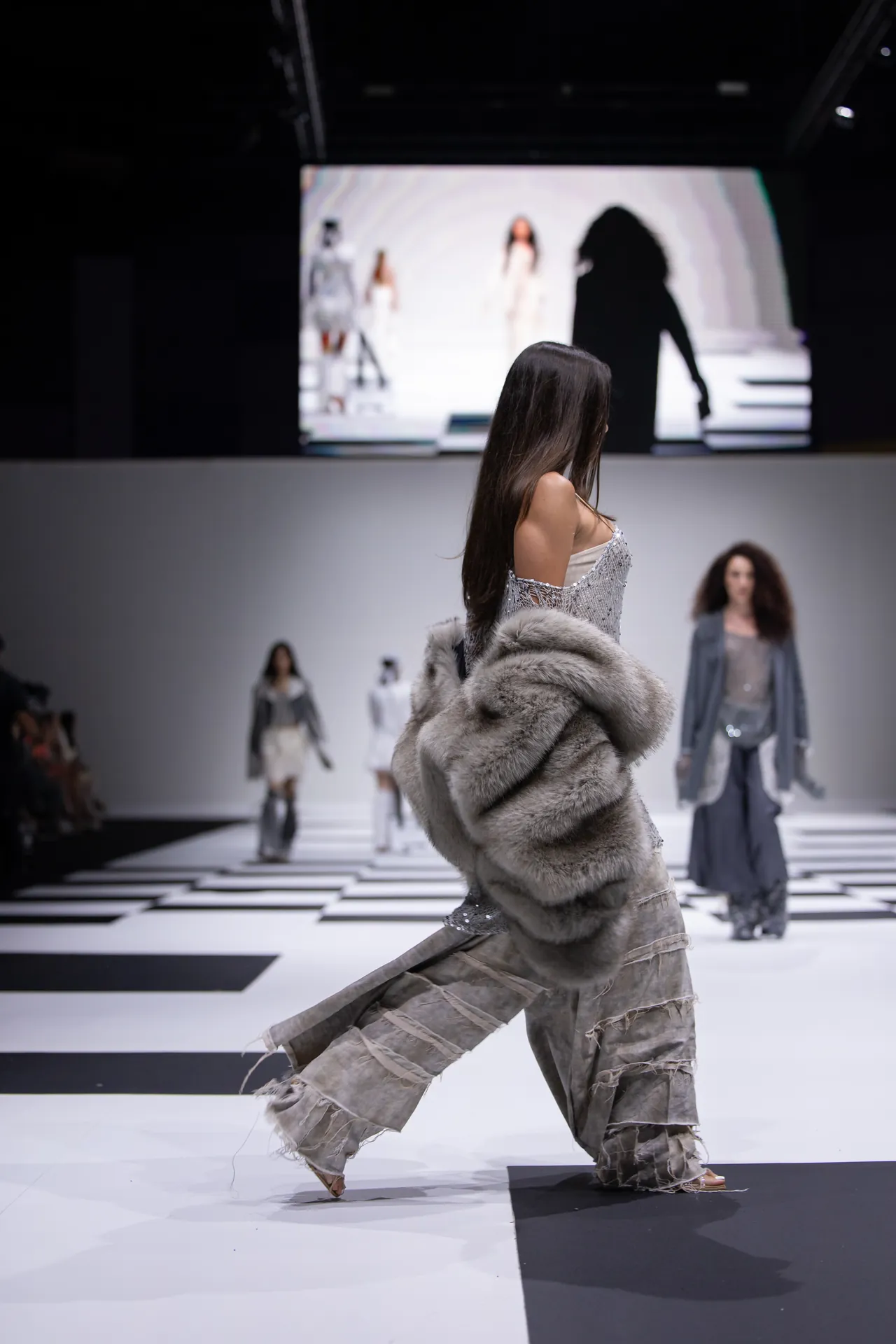 Model walking the runway in a long silver-gray fur coat with fringe, on a black-and-white checkered catwalk.
