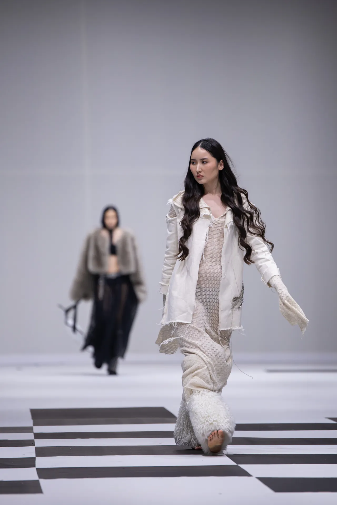 Model walks a runway in a cream textured dress with a white jacket on a black-and-white checkered catwalk; another model appears blurred in the background.