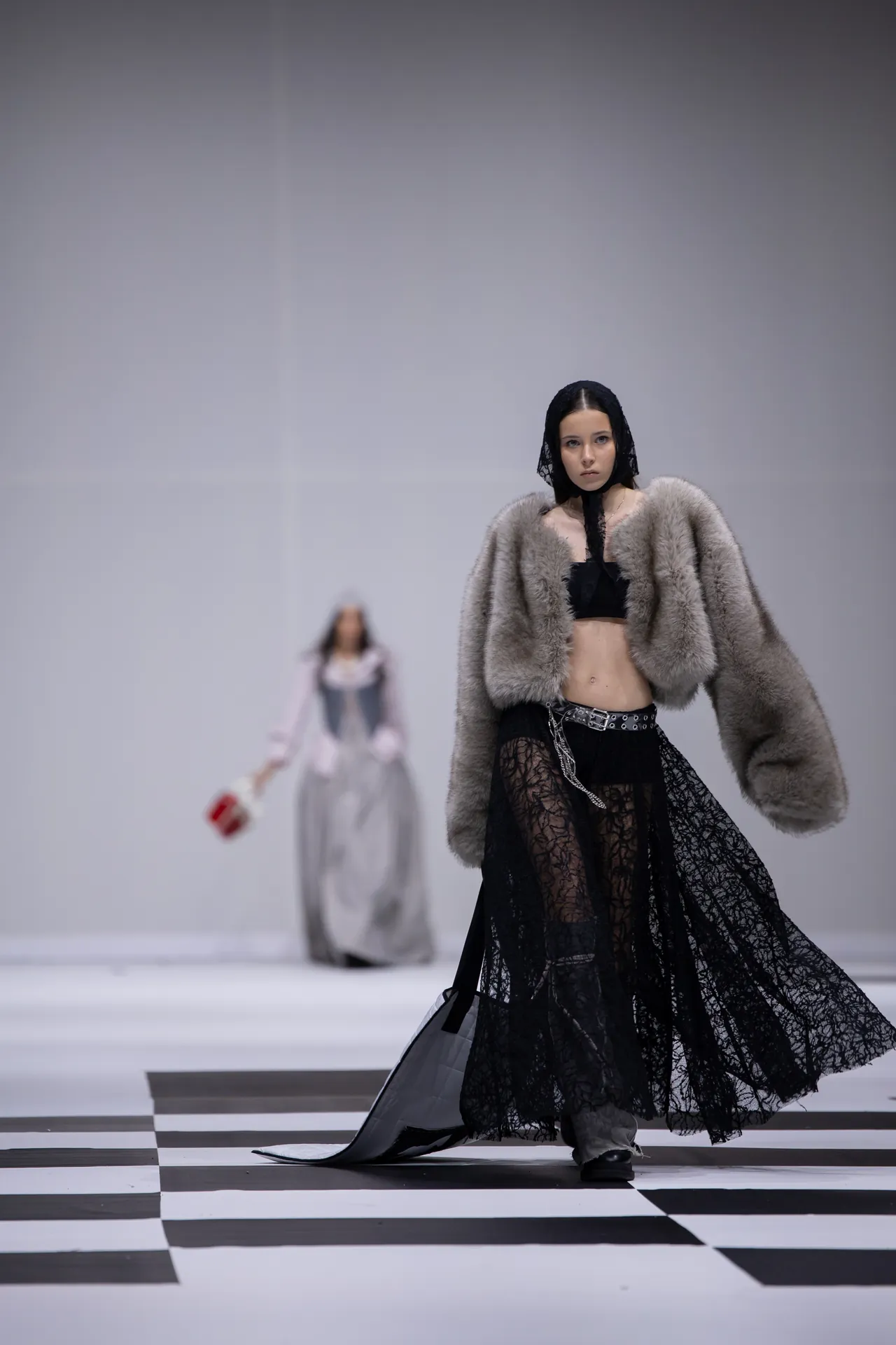Model walking a fashion runway in a black lace gown and gray fur coat on a black-and-white checkered floor.