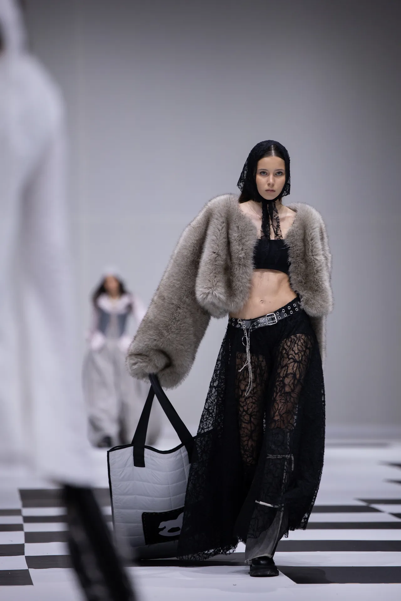 Model walking the runway in a plush fur coat over a black lace outfit, with a black headscarf and a white quilted tote bag.