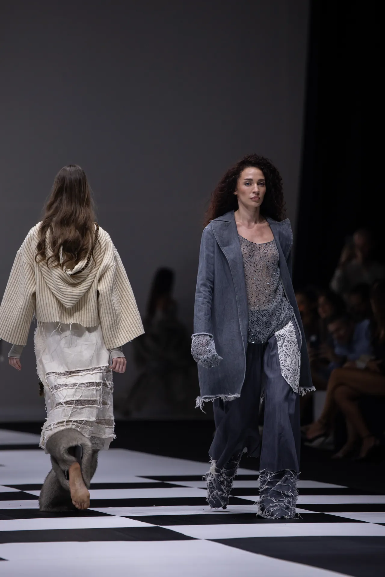 Two models walk a fashion runway, wearing distressed, layered outfits on a black-and-white checkerboard floor.