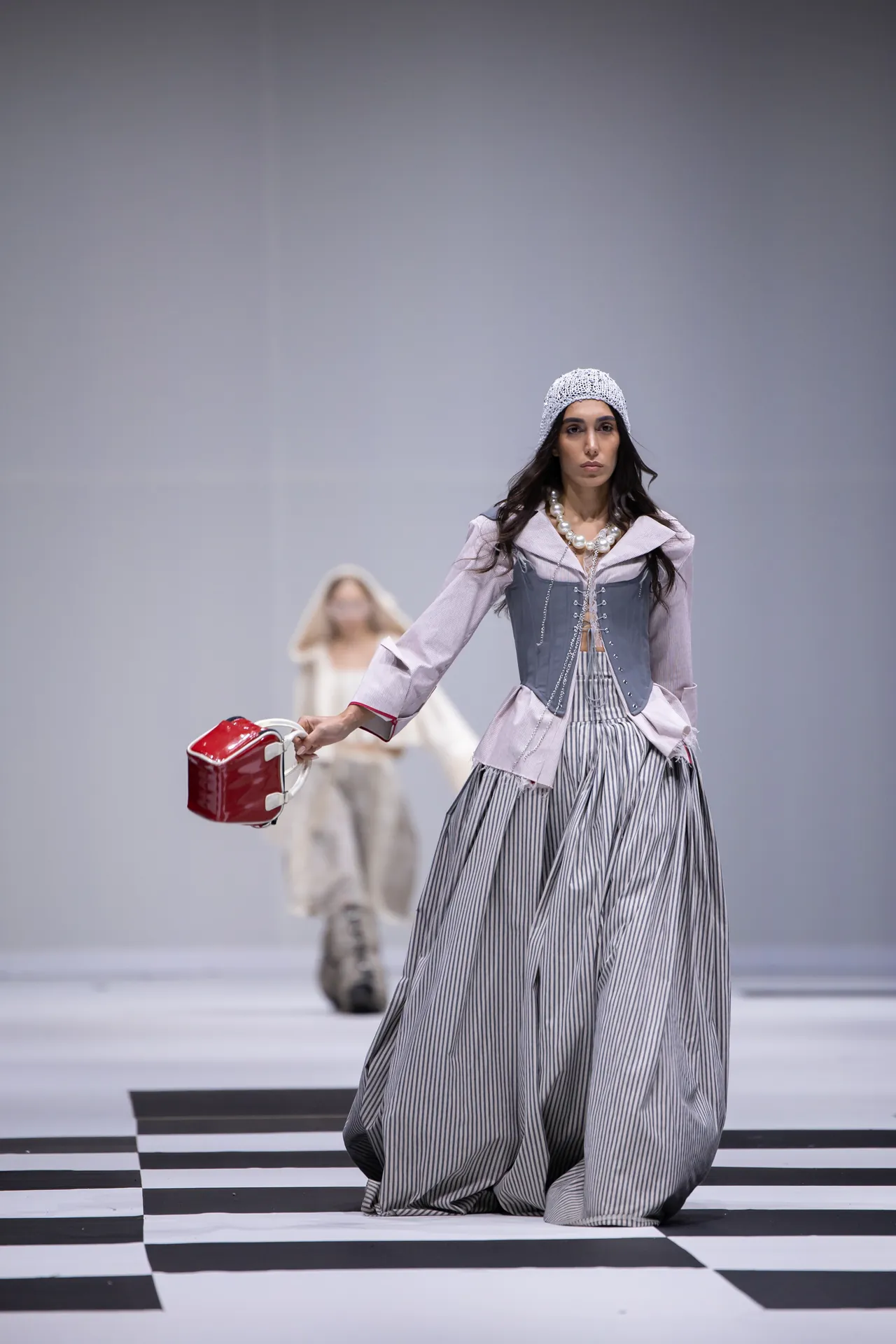 Model walks the runway in a pale pink-gray ensemble with a structured gray corset over a striped, voluminous skirt, pearl necklace, knit beanie, and a red handbag.