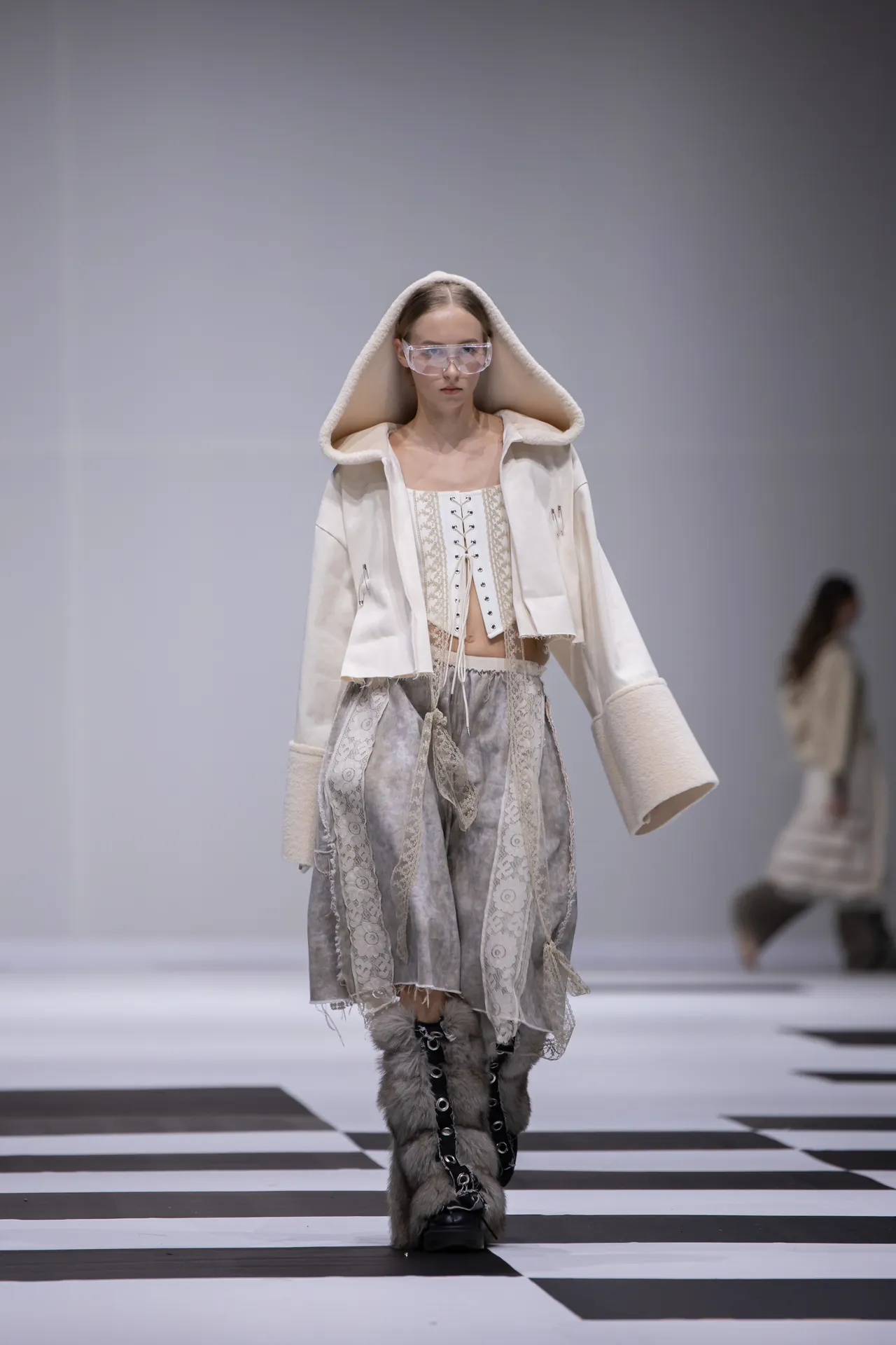 Runway model wearing a white hooded coat with fur trim, lace corset top, lace skirt, and furry boots on a black-and-white checkerboard catwalk.