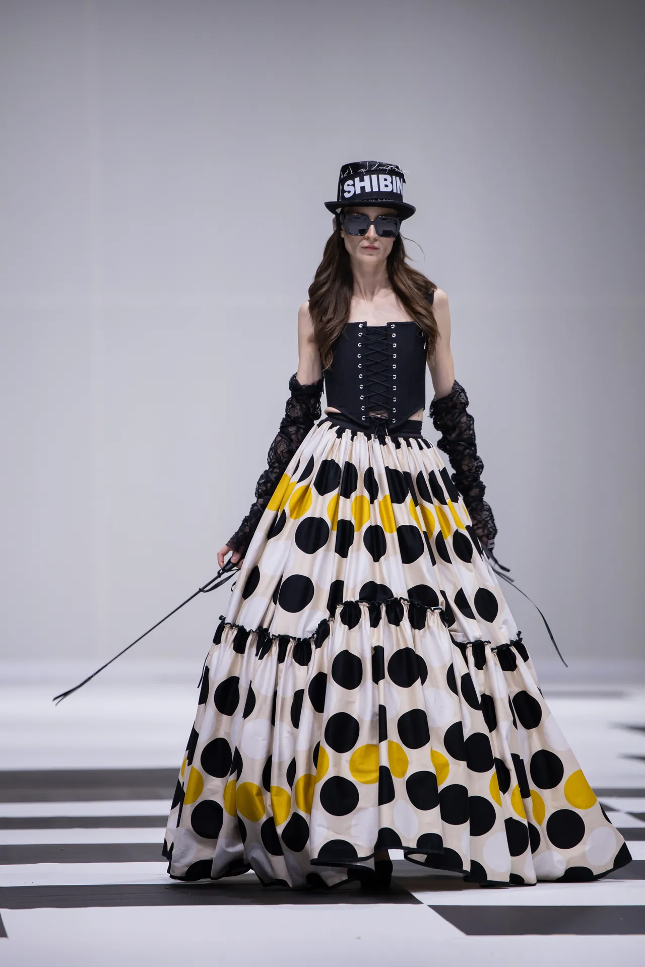 Model on a runway wearing a black corset top, dramatic polka-dot skirt with yellow accents, black lace gloves, sunglasses, and a cap reading SHIBIN.