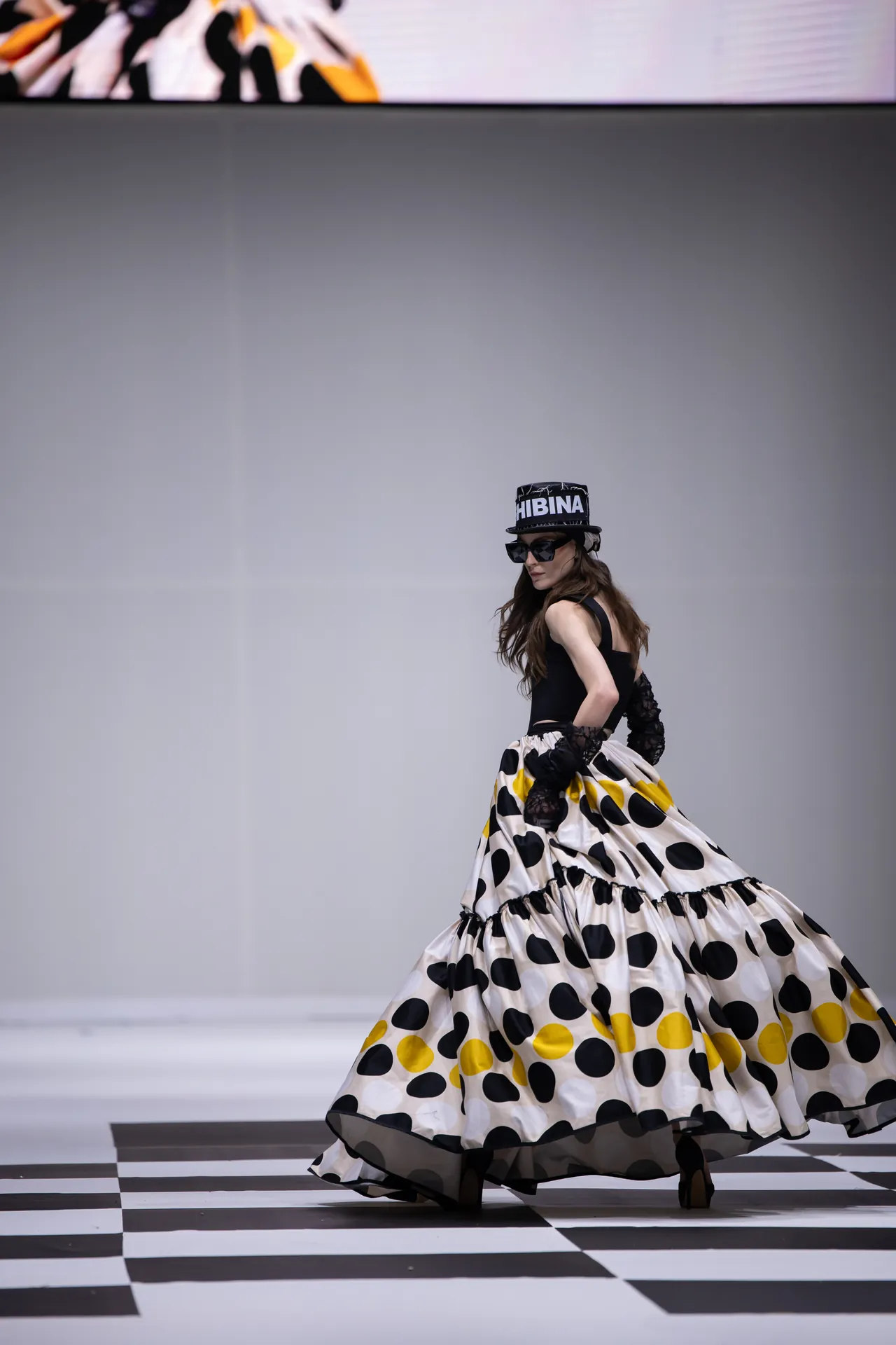 Runway moment: a model in a black sleeveless top and a voluminous polka-dot skirt walks across a black-and-white checkered floor, wearing gloves, sunglasses, and a hat with bold lettering.
