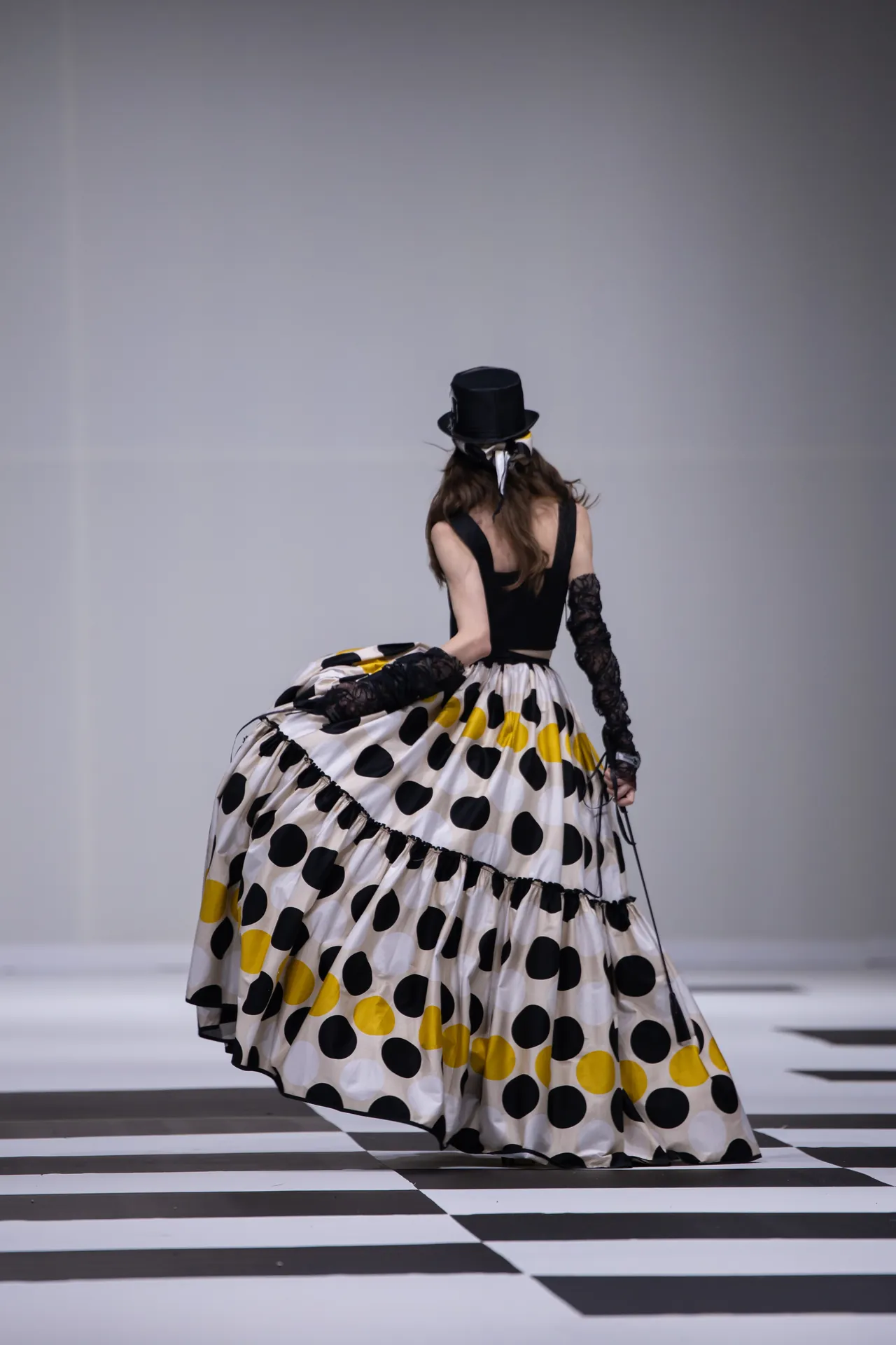 Back view of a model walking a fashion runway in a black bodice, large white skirt with black and yellow polka dots, lace gloves, and a top hat on a striped floor.
