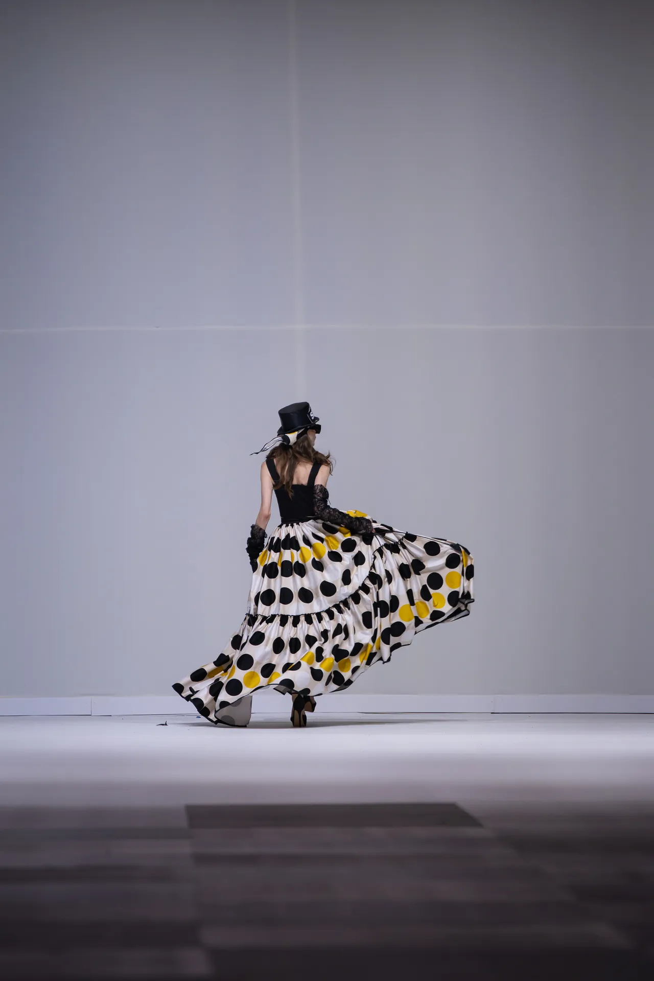 A woman in a black top hat and a long flowing polka-dot skirt twirls on a runway or stage, back to camera.