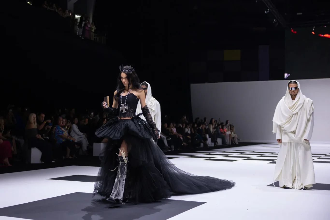 Model in an elaborate black gown with a crown walks the runway; audience sits along dark backdrop at a fashion show.