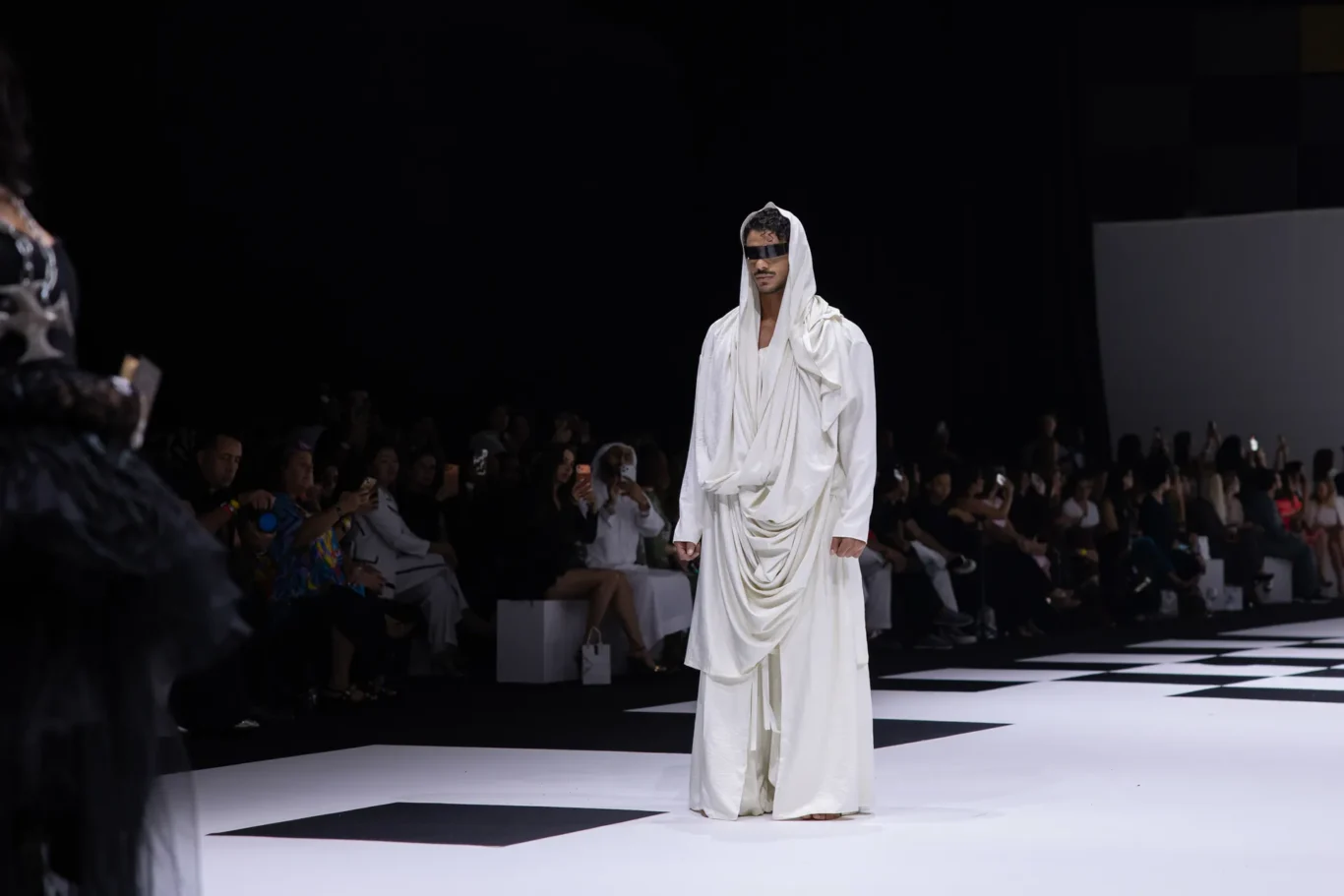 Model in a flowing white robe with a hood and black visor sunglasses walks a black-and-white checker runway while spectators watch from the sides.