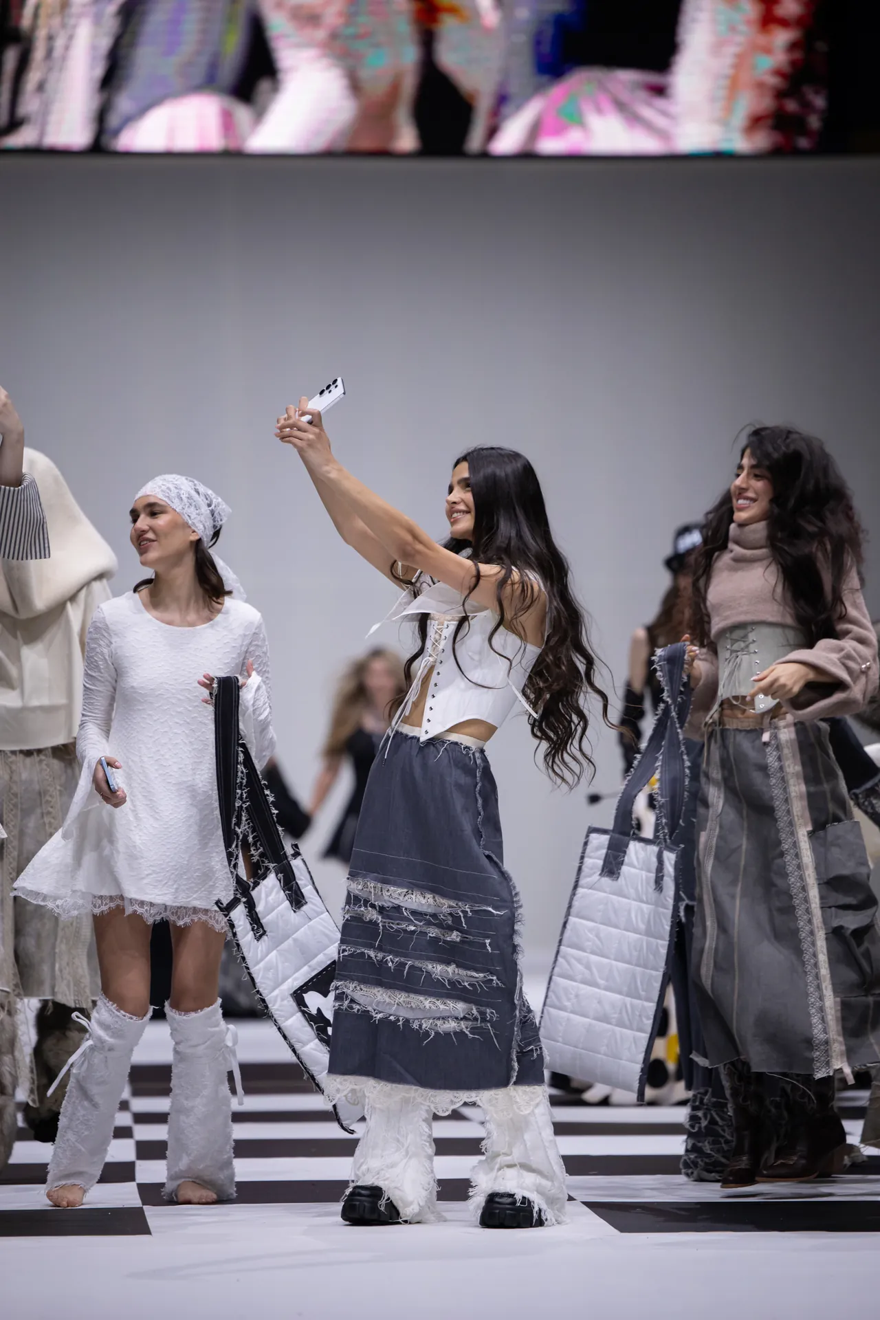 Model on a fashion runway raises a phone to take a selfie while other models pose with white quilted bags nearby.