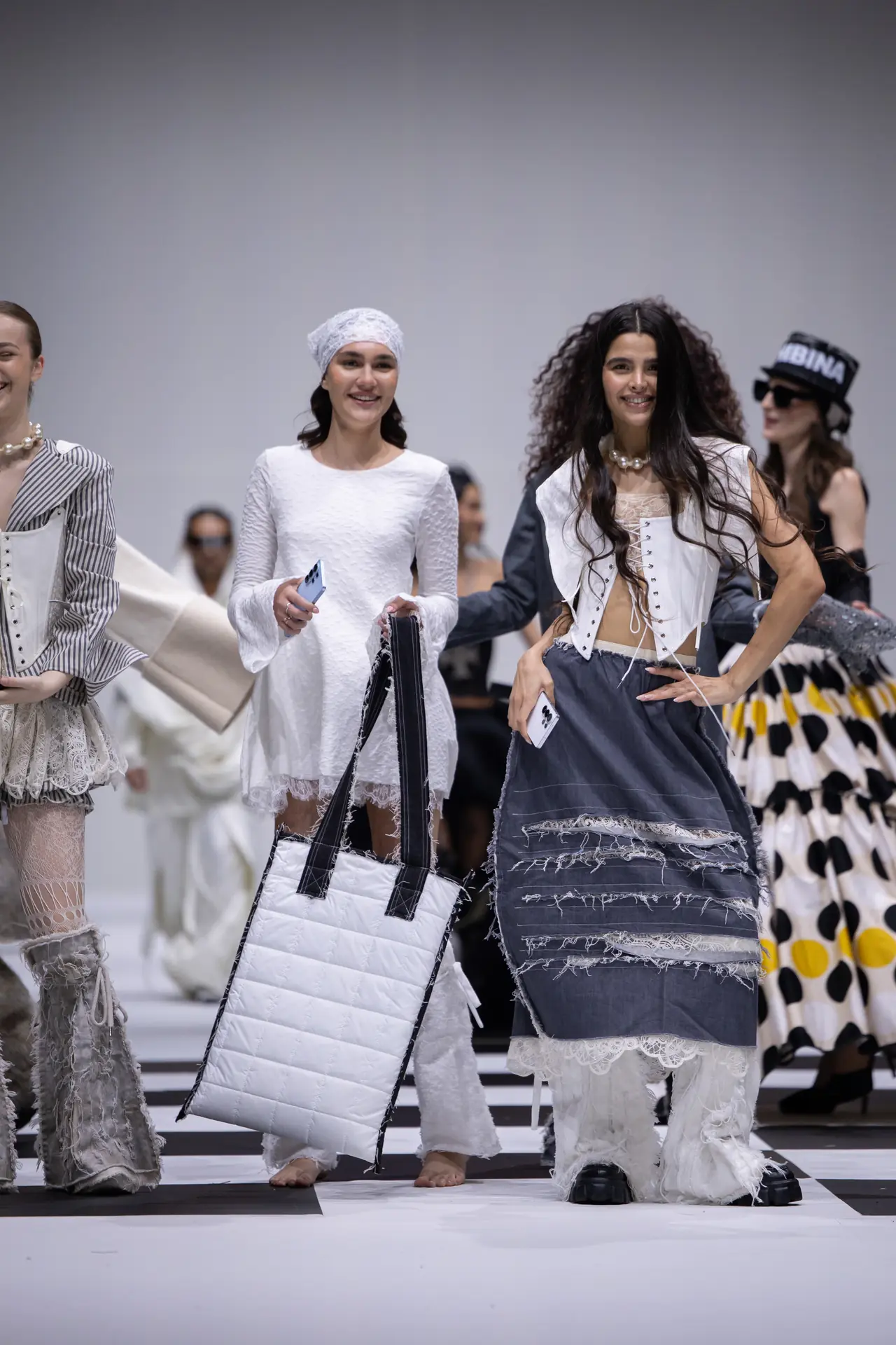 Models walk a fashion show runway in white and denim outfits, each holding large quilted bags.
