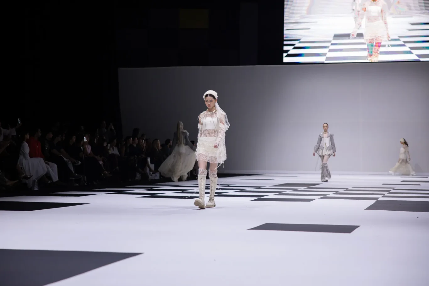 Model wearing a white lace outfit and thigh-high boots walks the runway as the audience sits along the left side of the catwalk, dimly lit backdrop behind.