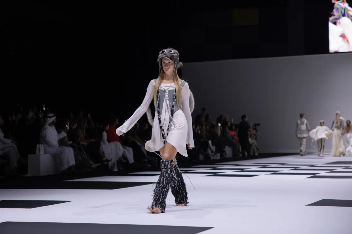 Model walks the runway in a gray-white avant-garde outfit with fur accents on a black-and-white checkered catwalk surface.