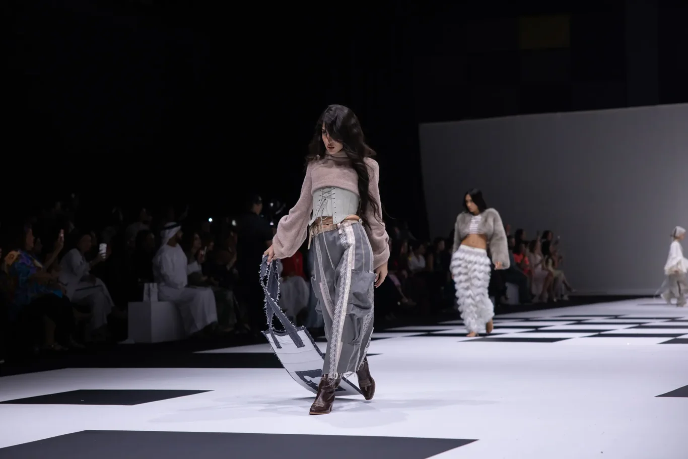 Model walking a fashion runway in a lavender sweater with a corseted gray top, gray pants, brown boots, and a large white tote bag; catwalk and audience in dim backdrop nearby.