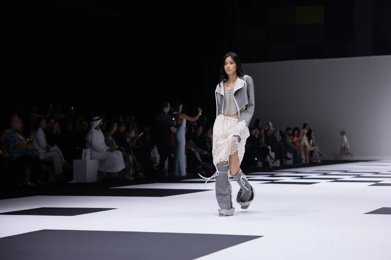 Model walks a black-and-white checkered runway in a gray lace-up outfit with a corset, audience along the sides.