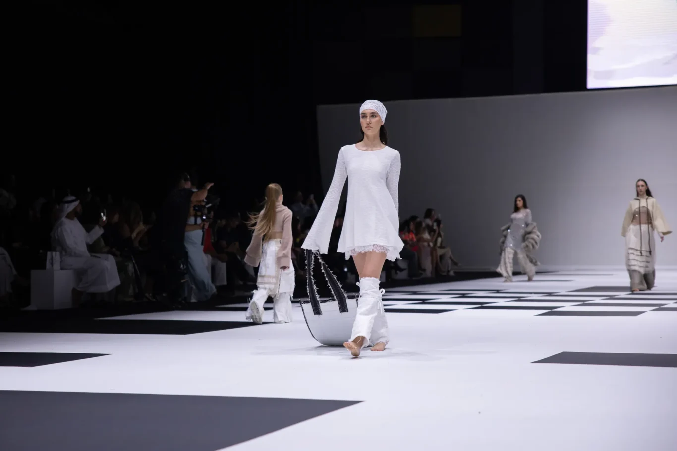 Model in a white textured dress and headscarf walks the runway on a black-and-white geometric stage.