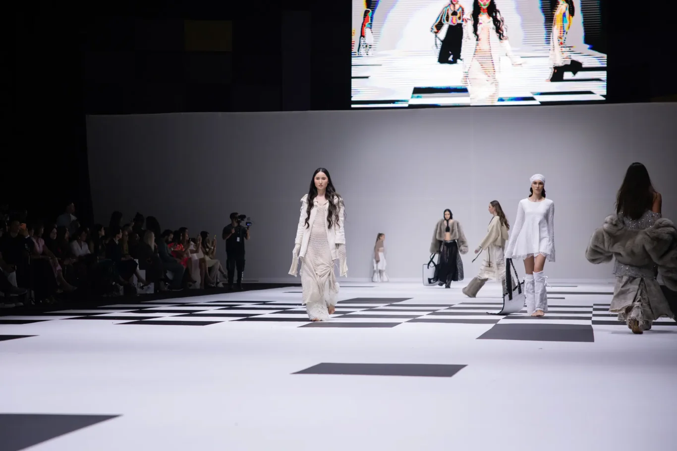 Models walk a fashion runway in cream and white outfits as spectators watch from the left.