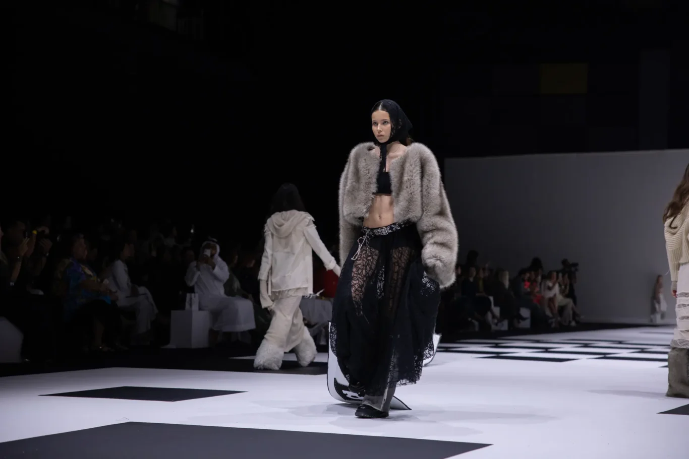 Model on a fashion runway wearing a gray fur coat over a black lace outfit, with a headscarf and chain belt.