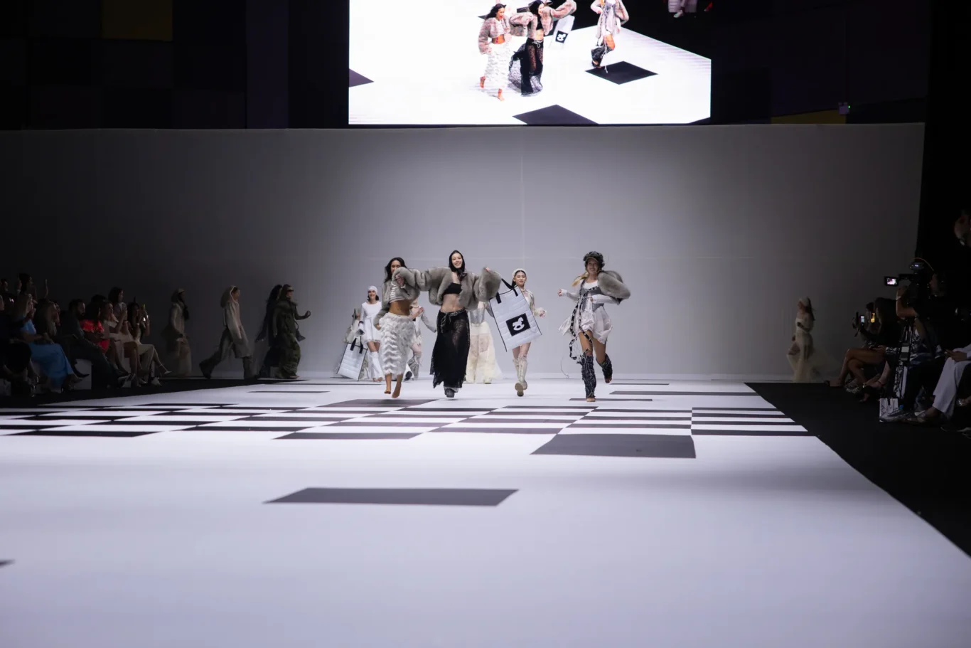 Models walk a black-and-white geometric runway as the audience watches from both sides during a fashion show.